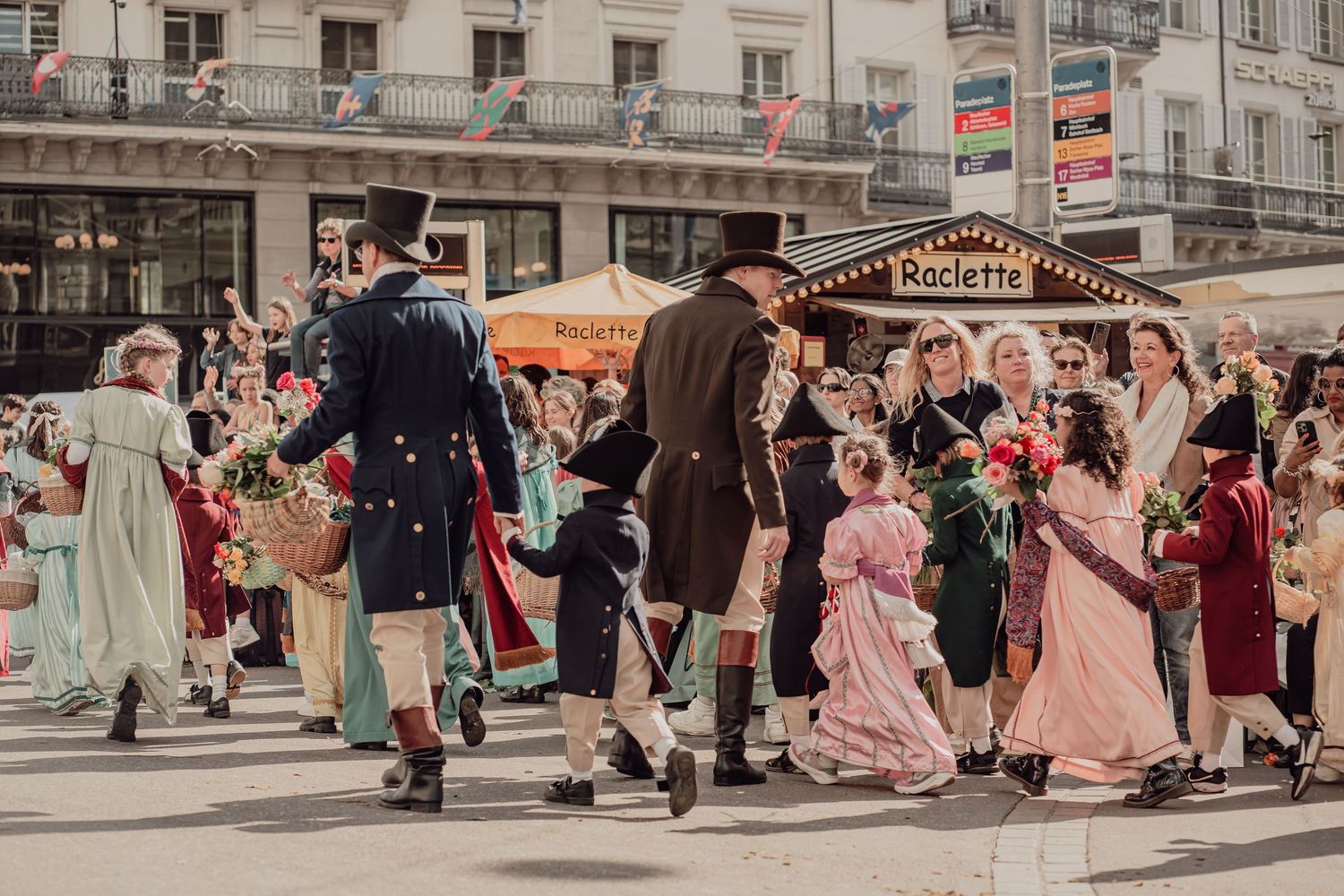 People in Victorian-era costumes dancing and parading through a festive town square with flowers.