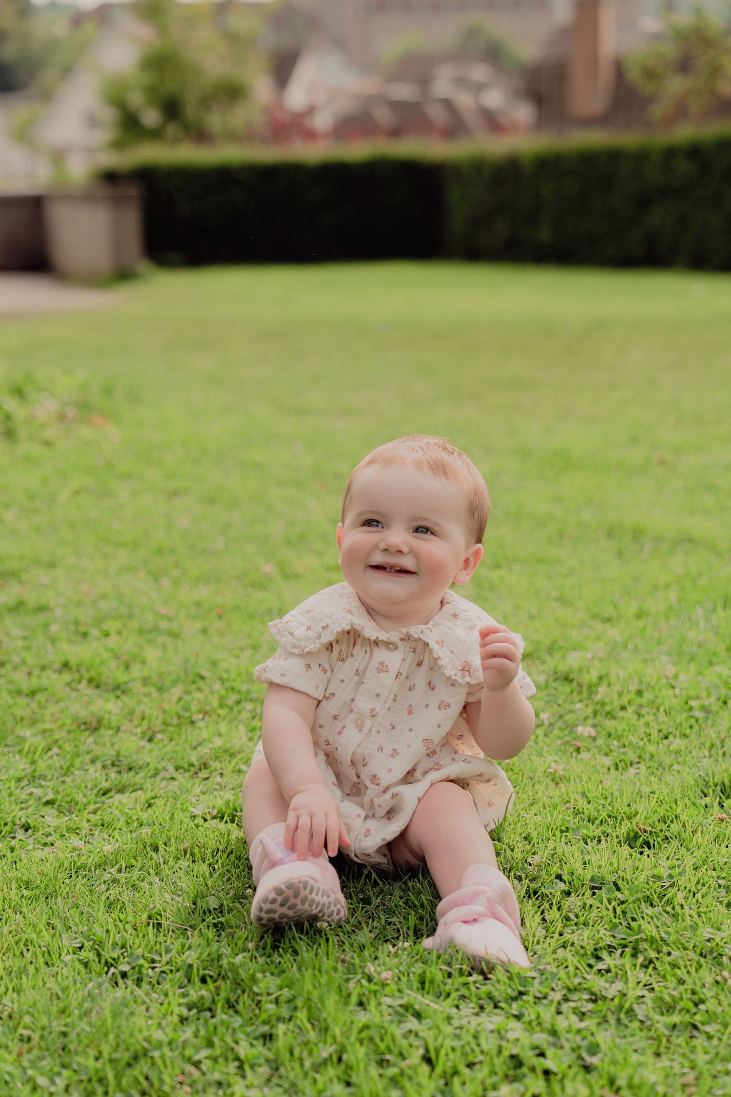 Happy baby in floral romper smiling while sitting on lush green grass in garden setting.