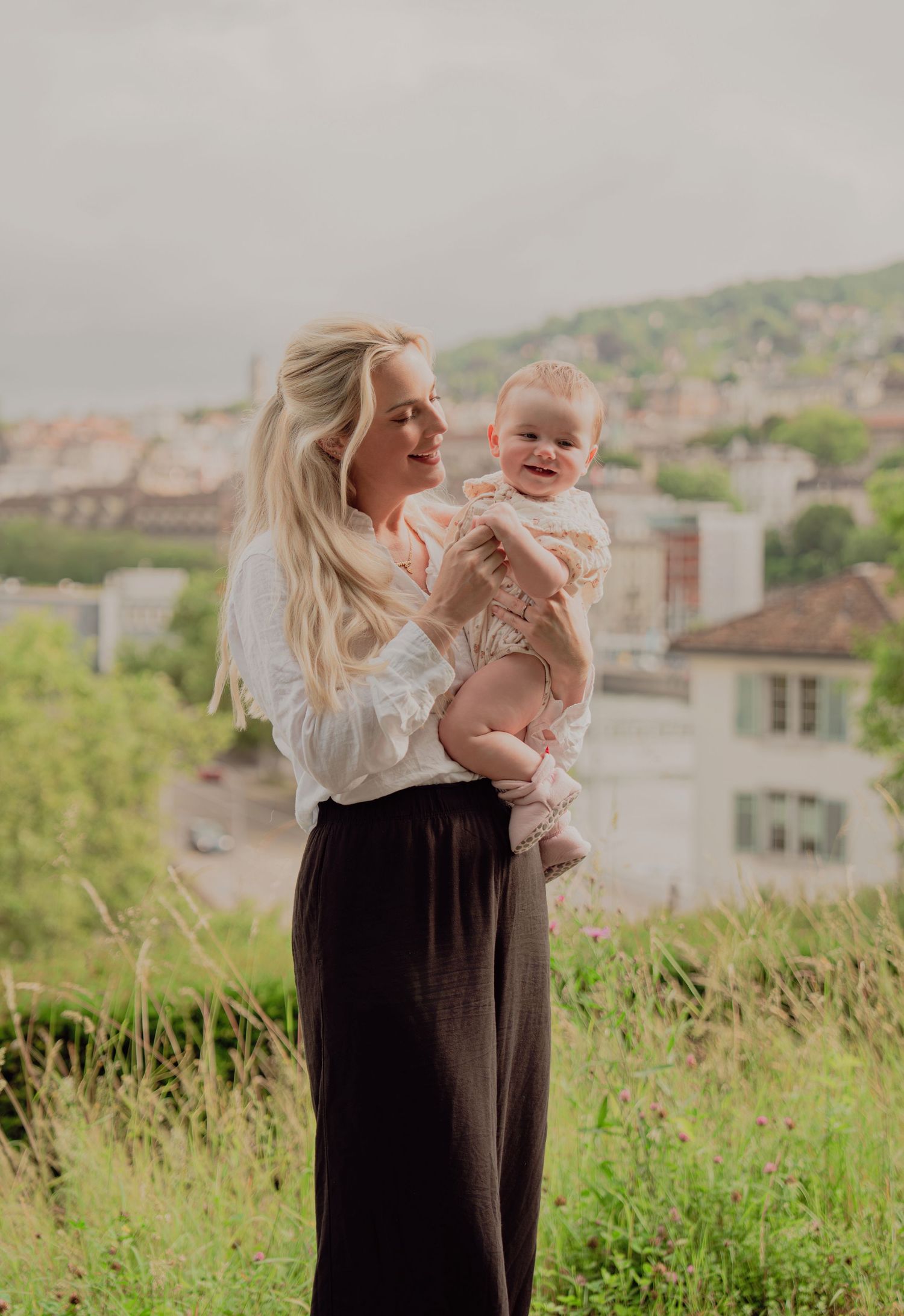 Blonde mother holding smiling baby on a grassy hilltop with a European cityscape in background.