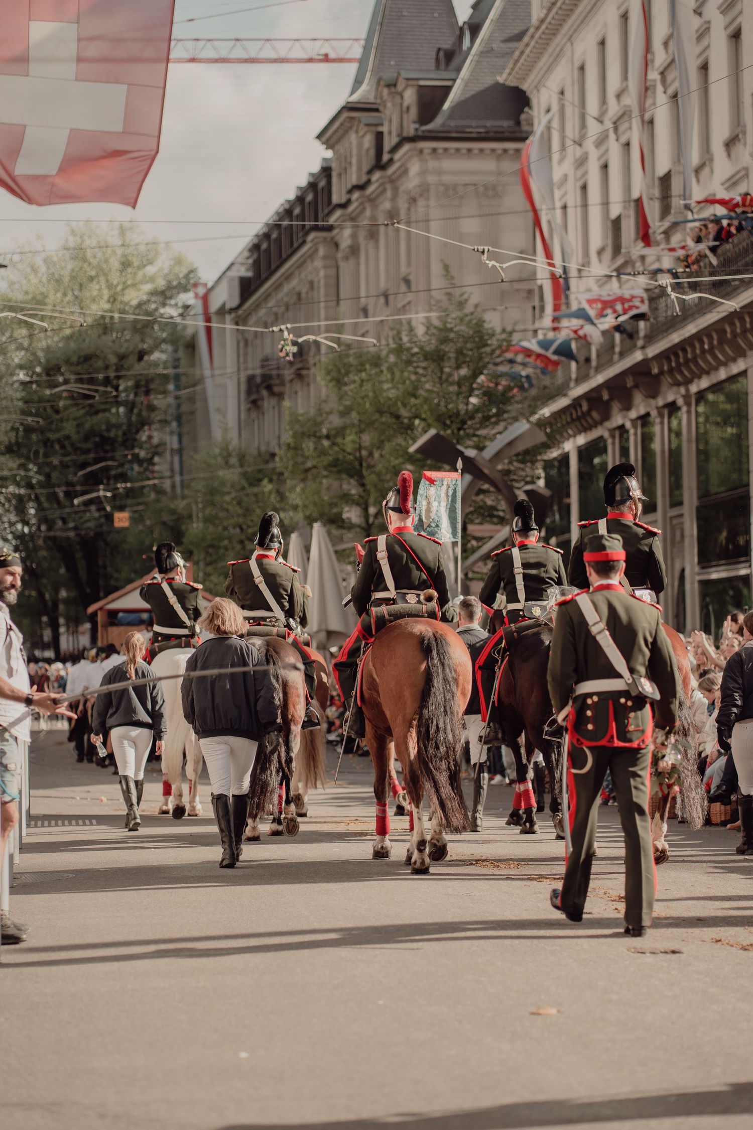 Riders in historical military uniforms on horseback parade down a city street under Swiss flags.