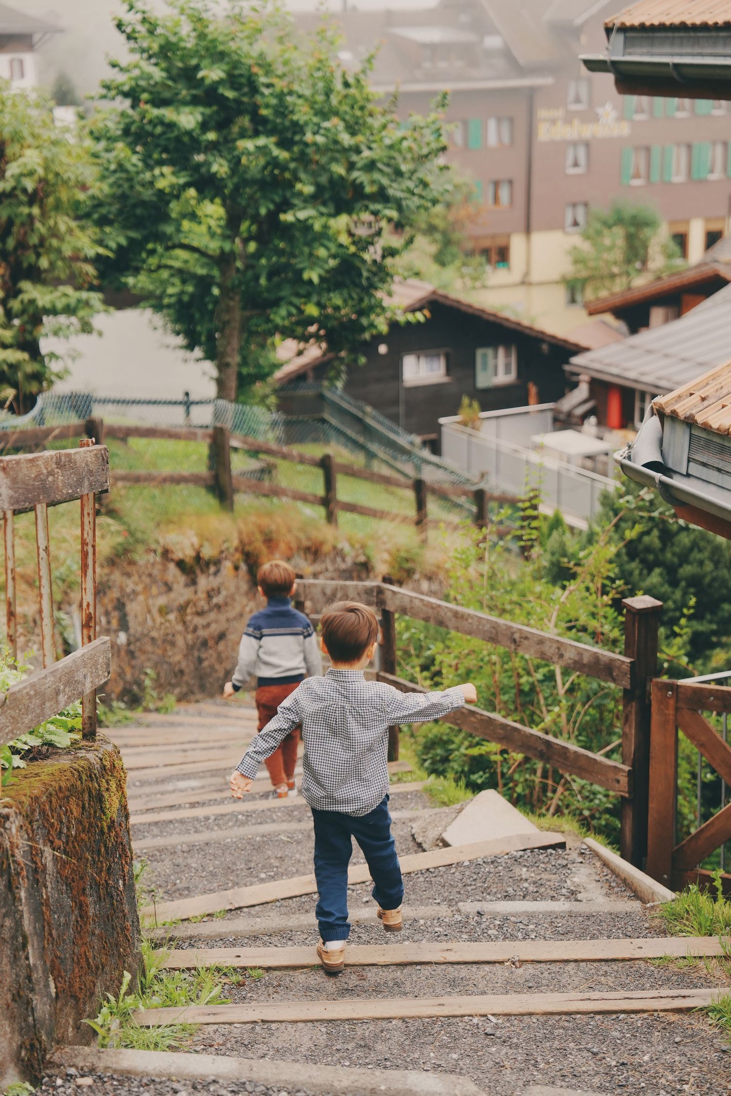 Two young boys walk up rustic wooden outdoor stairs in a scenic alpine village with houses.