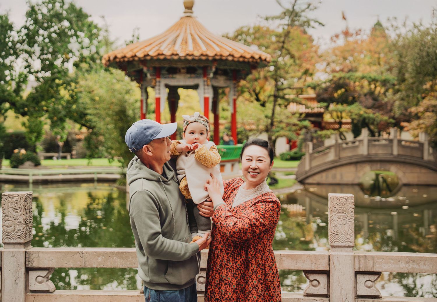 Couple holding baby girl laughing together near ornate Chinese gazebo and pond in a lush garden setting.