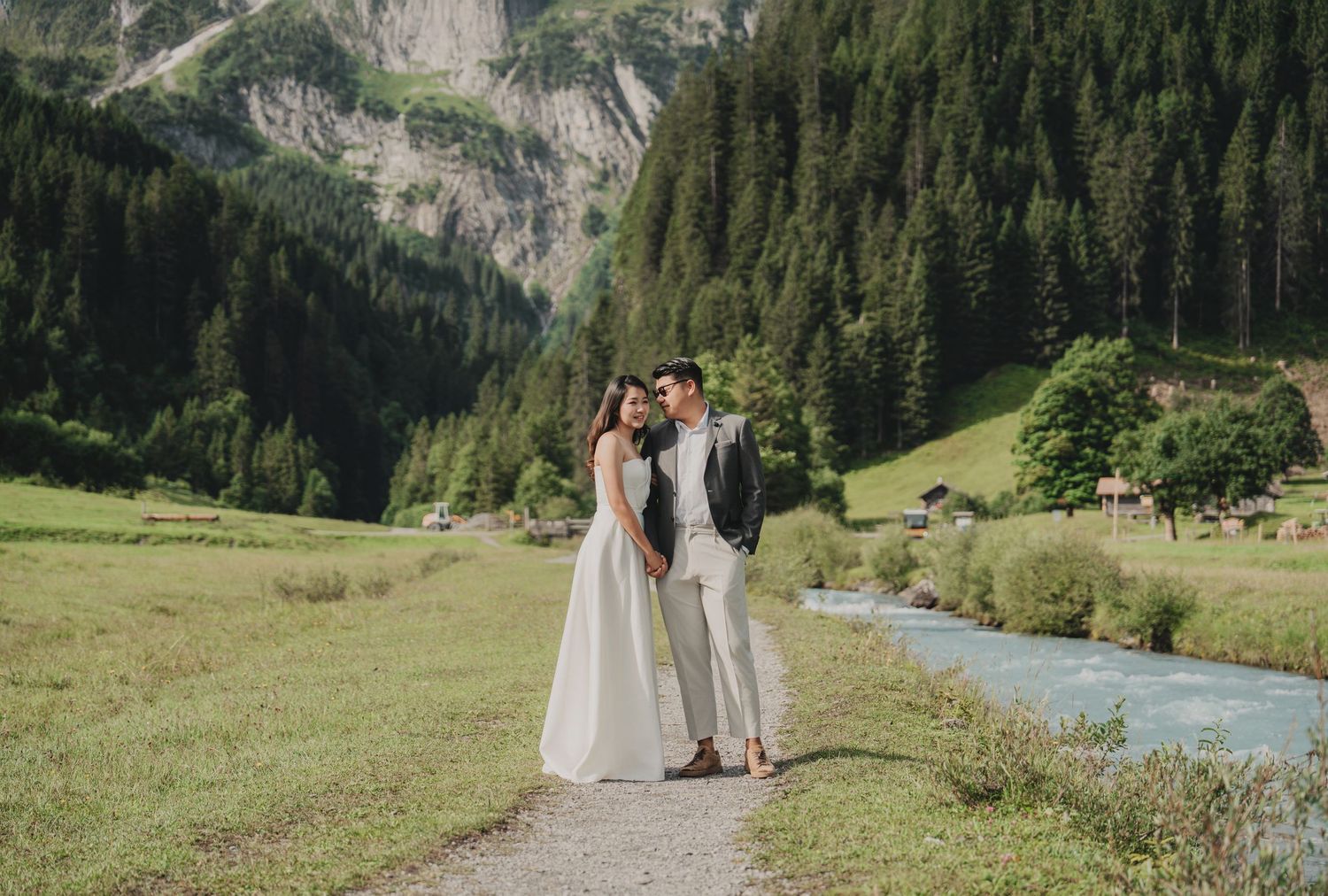 Wedding couple standing on a path by a glacial stream surrounded by lush Alpine meadows.
