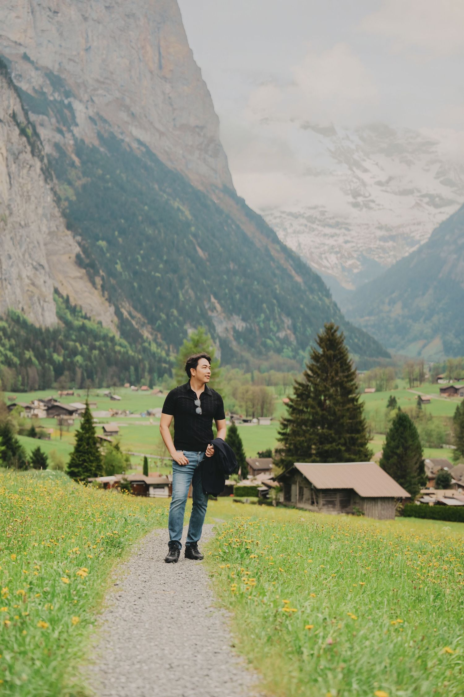 Young man walking on path through lush Swiss valley with dramatic snow-capped mountains and green meadows.