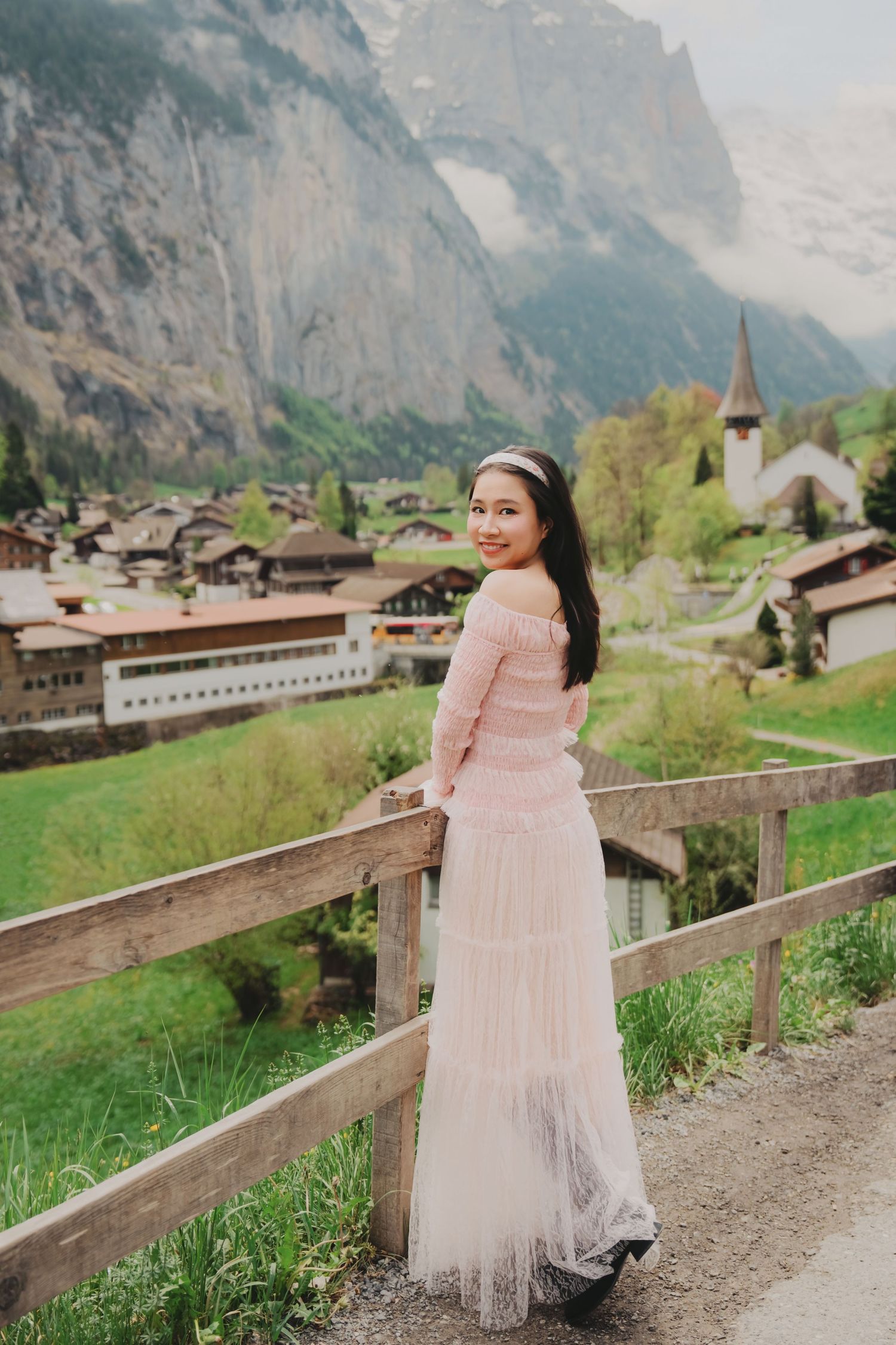 Smiling woman in elegant pink off-shoulder dress posing by wooden fence overlooking charming Swiss mountain village.