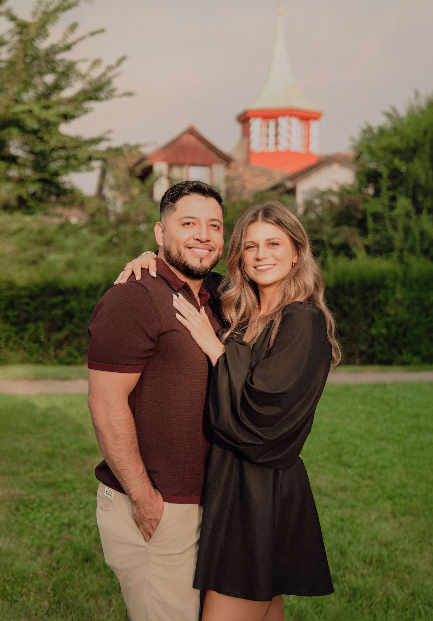 Smiling couple posing together in a green park with a charming red-roofed gazebo structure behind them.