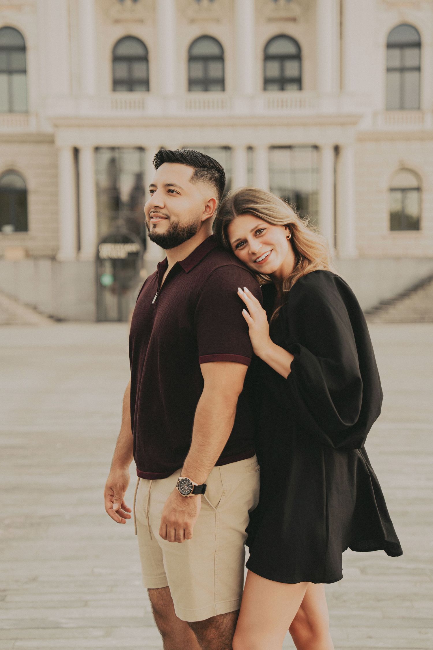 Woman in black dress leaning on mans shoulder as they pose back to back near a European building.
