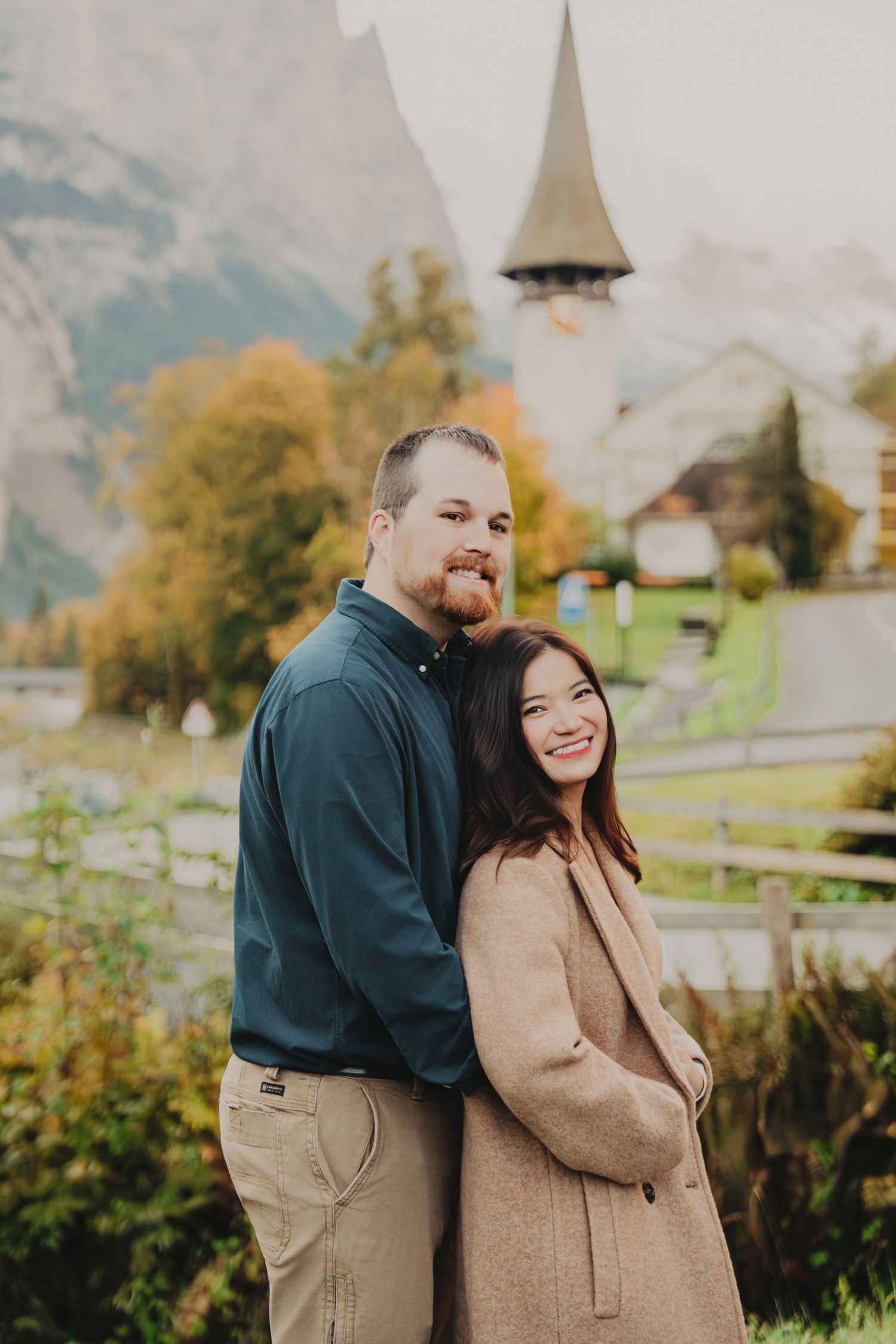 Couple smiling together with a Swiss church steeple and misty Alpine mountains in the autumn background.
