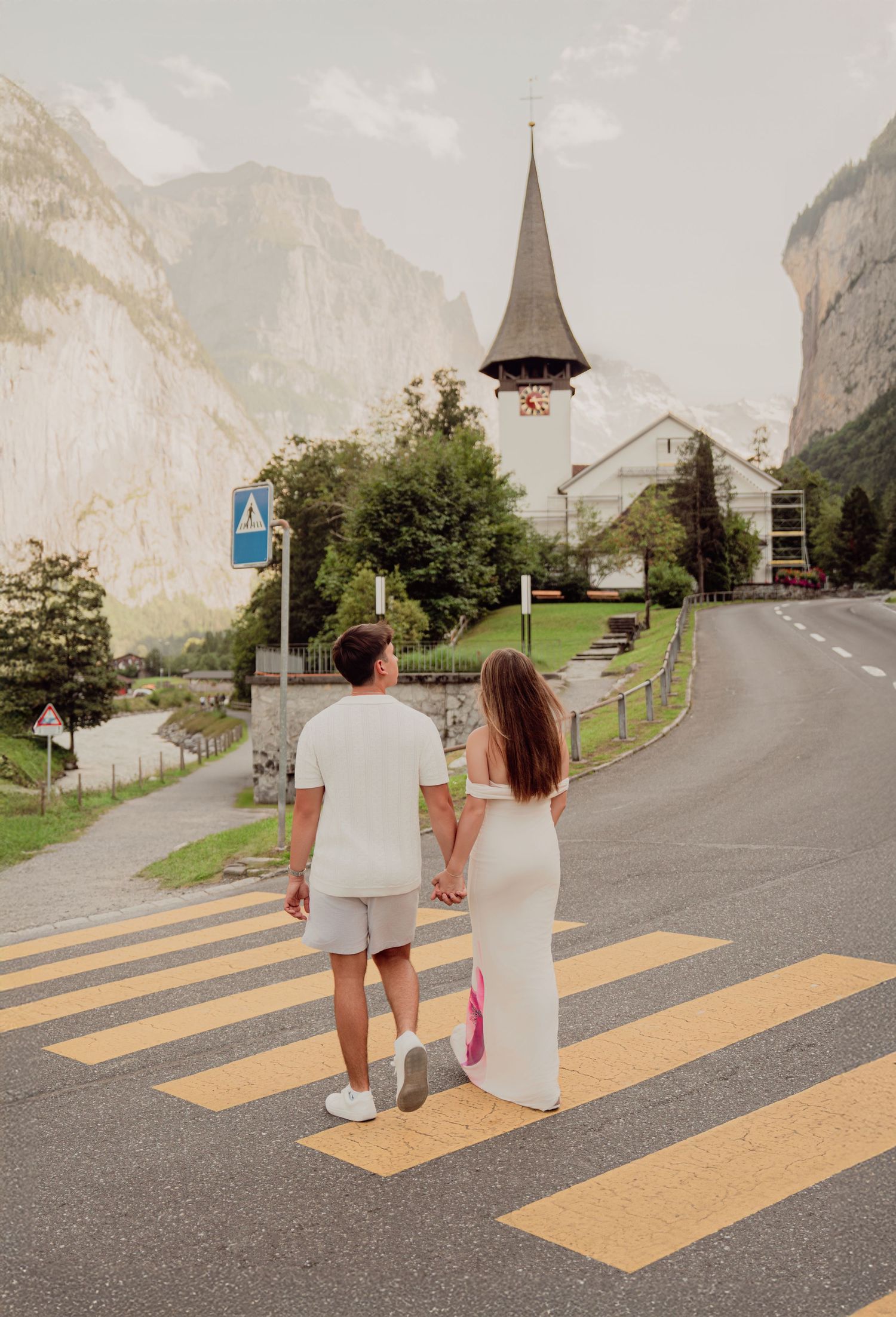 Couple holding hands crossing a zebra crossing with a charming Swiss church steeple and mountain valley backdrop.