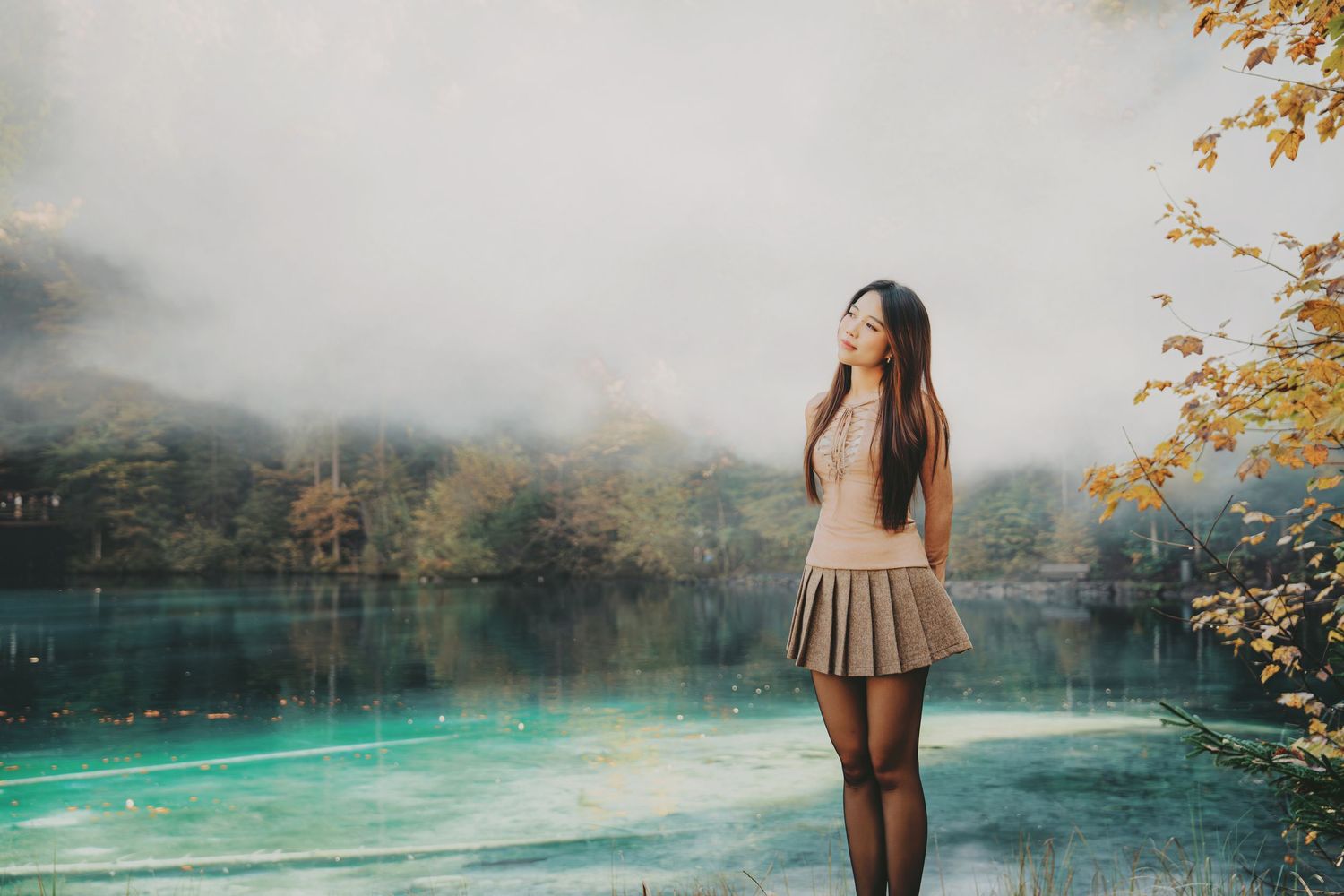Young woman in mini skirt standing by a misty turquoise lake with autumn trees behind her.
