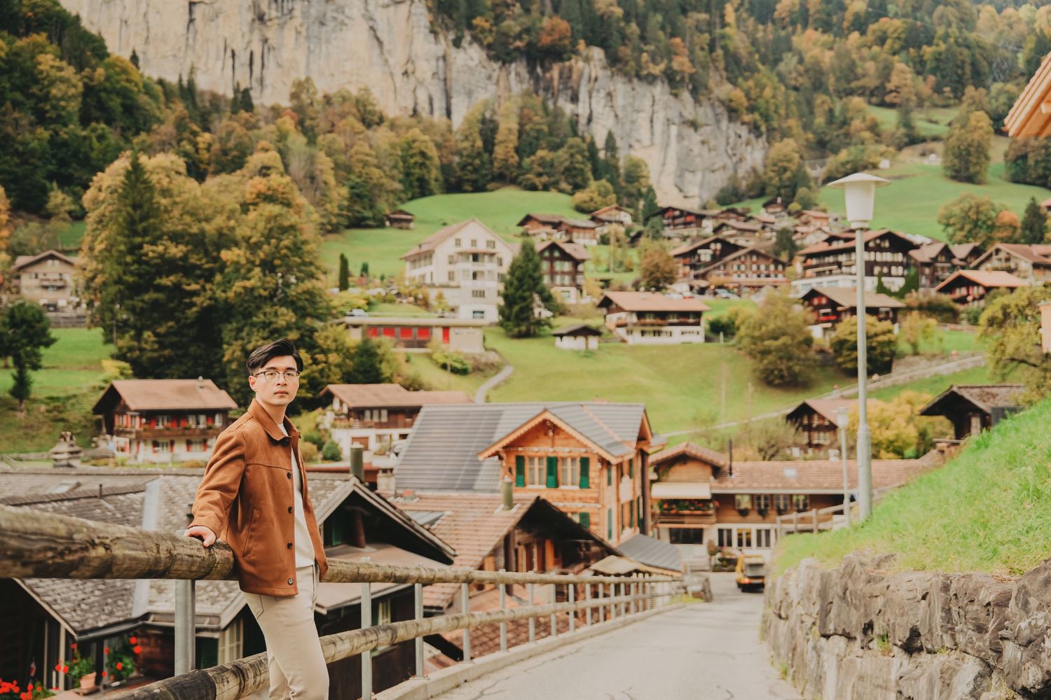 Man in brown jacket posing on bridge in Swiss alpine village Lauterbrunnen with traditional chalets and cliffs.
