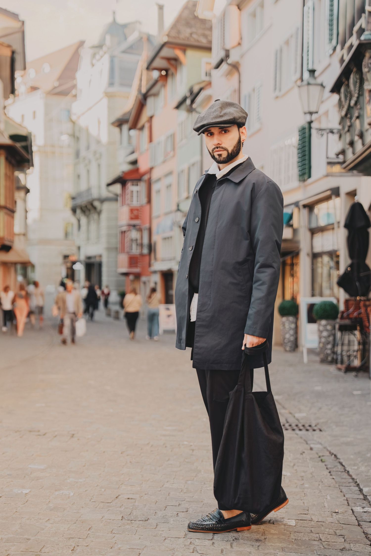 Stylish bearded man in grey coat and flat cap holding a tote bag on a cobblestone European street.