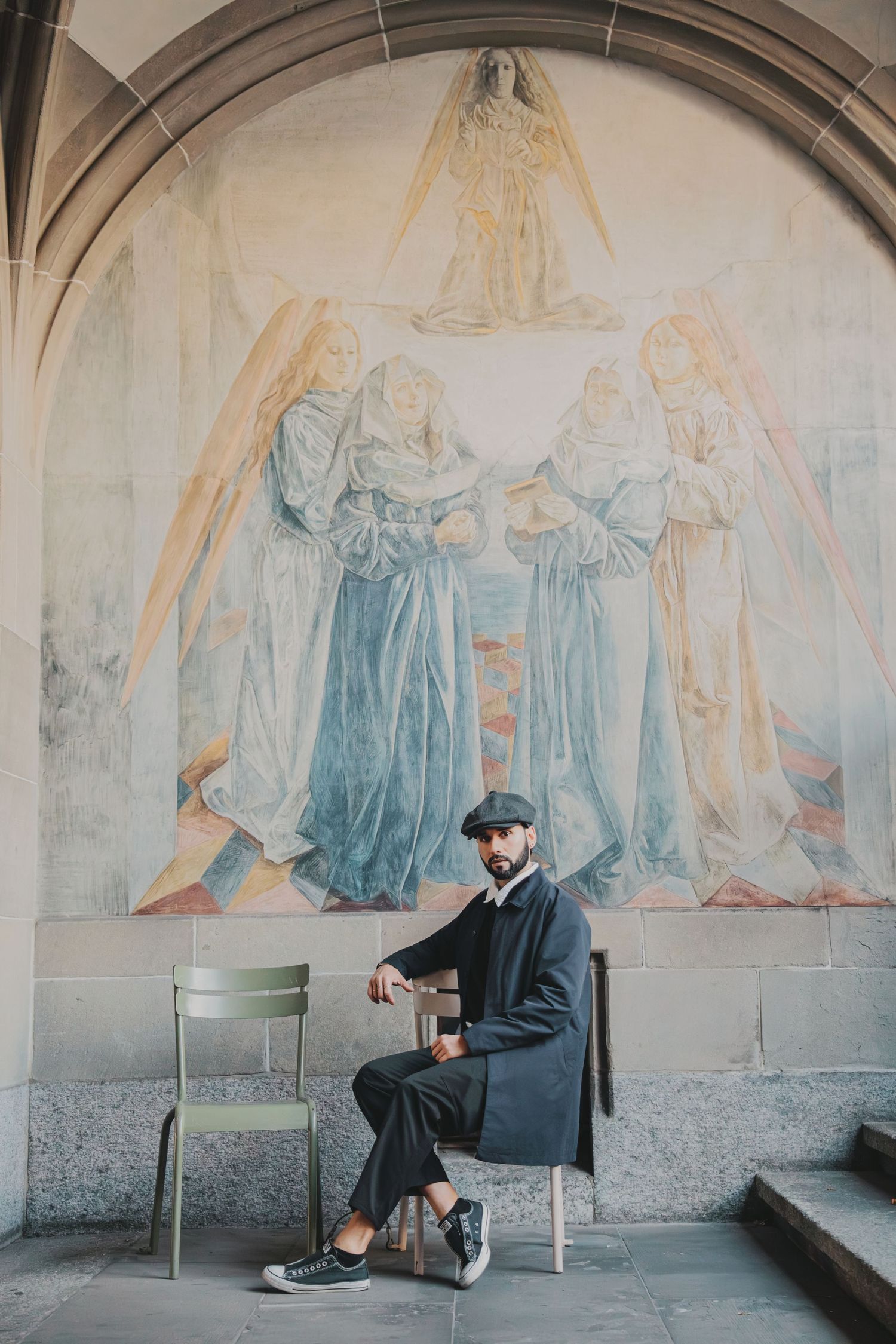 Stylish man in flat cap seated before ornate religious fresco painting in historic arched corridor.