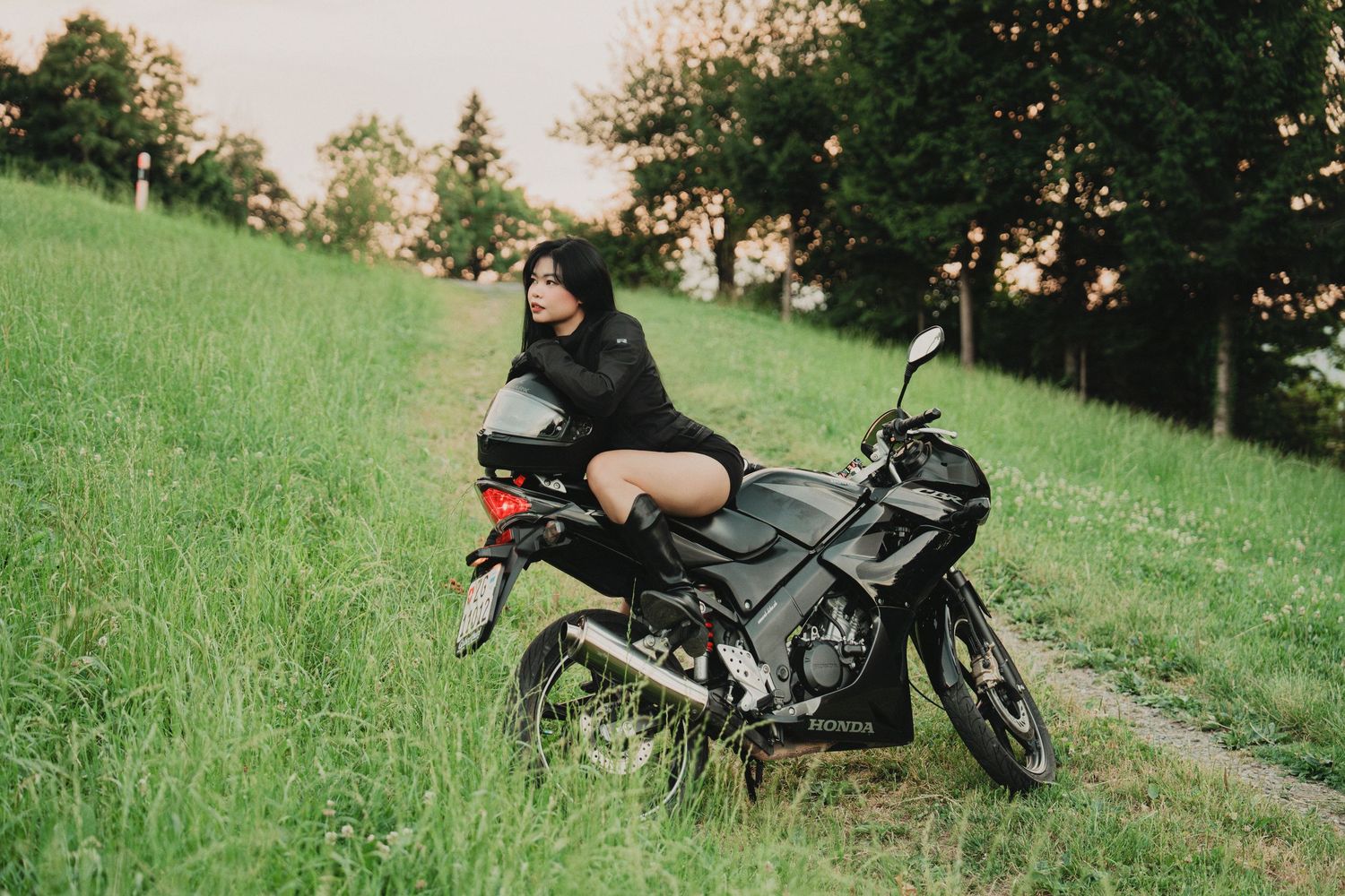 Woman in black outfit sitting on black sport bike on lush green hillside at dusk.