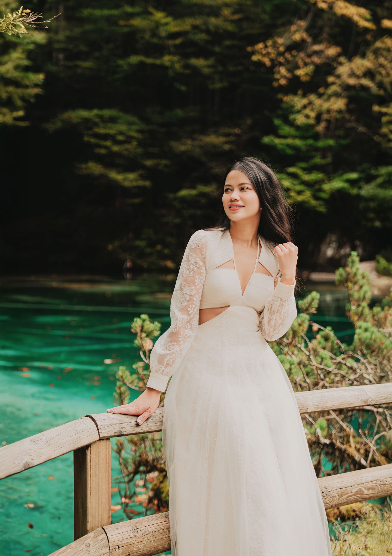 Smiling young woman in white lace crop top and skirt posing by wooden fence near turquoise lake.
