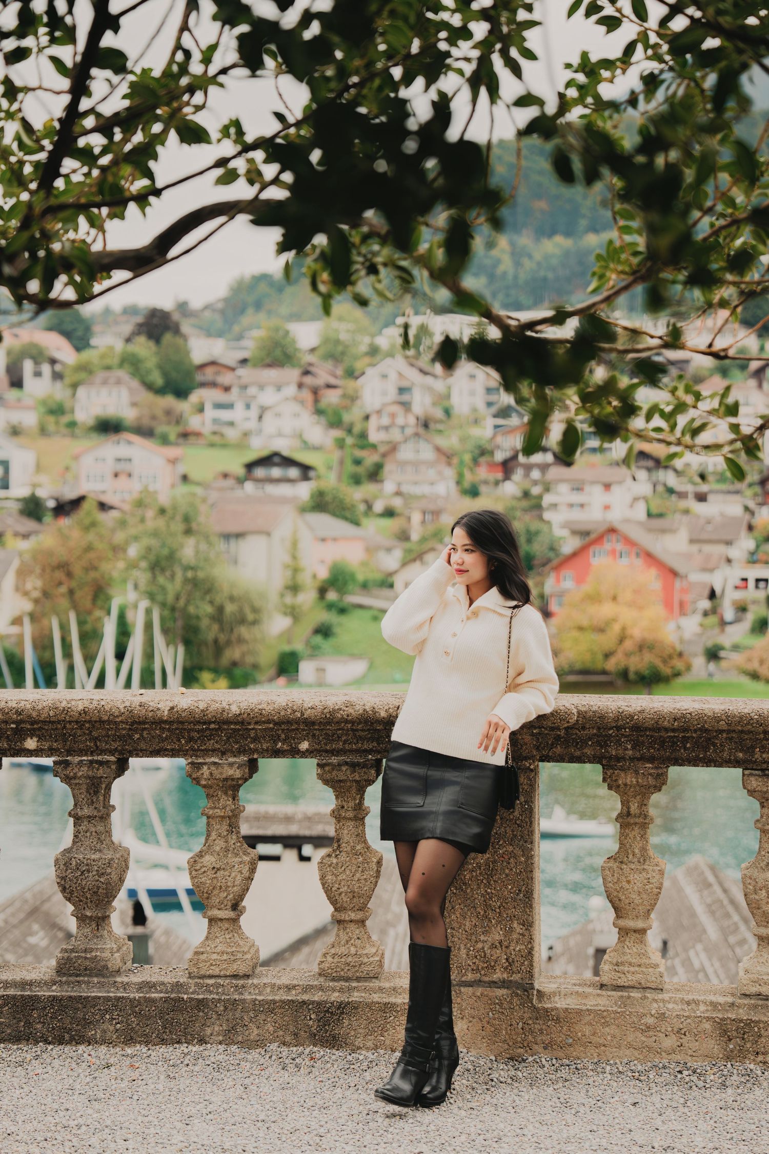 Young woman in cream sweater and black mini skirt poses by stone balustrade overlooking Swiss village.