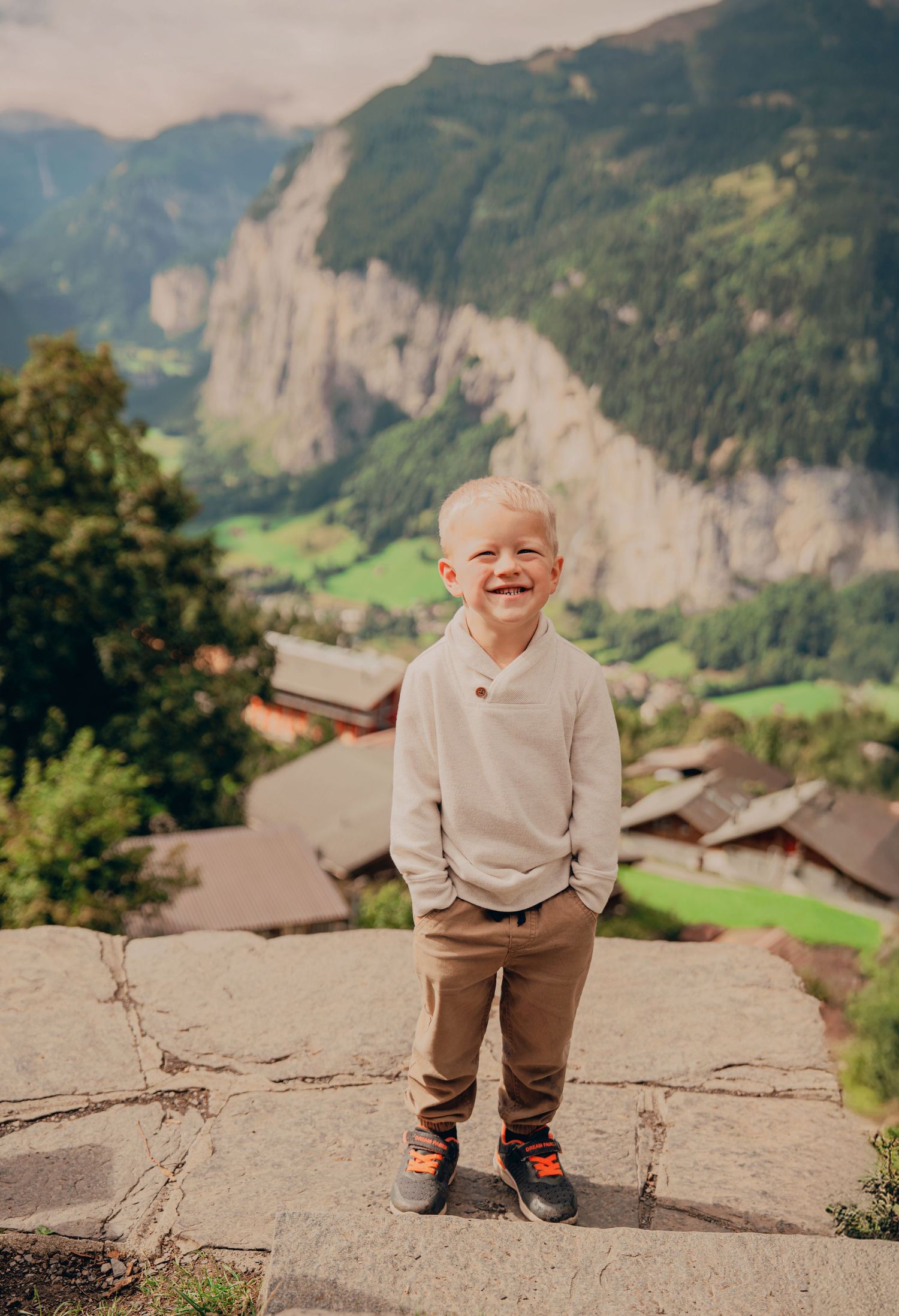 Joyful young boy in cream sweater and brown pants laughing on stone wall with Alpine scenery behind.