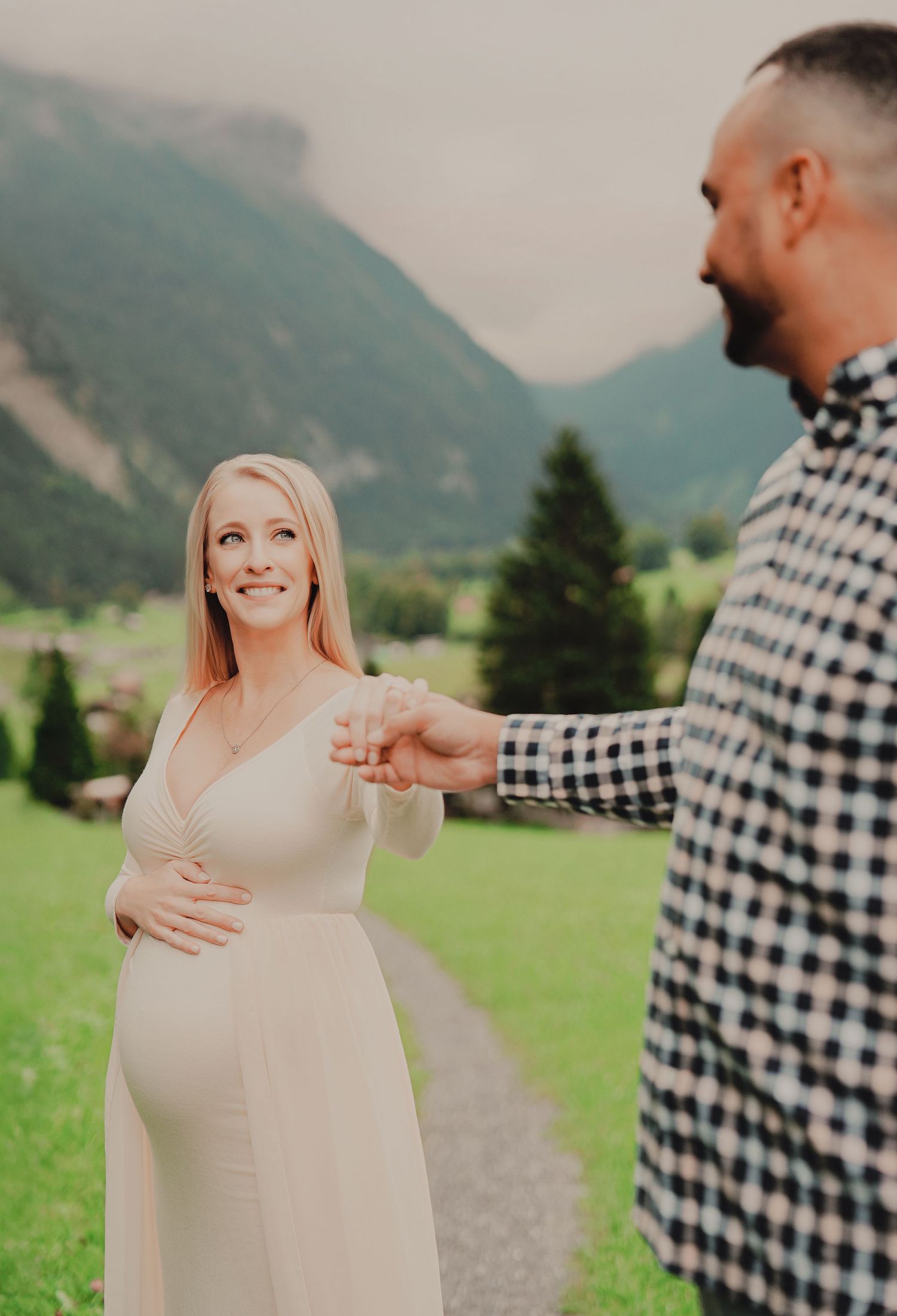Smiling pregnant woman in white dress holding hands with her partner on a scenic Swiss mountain meadow path.
