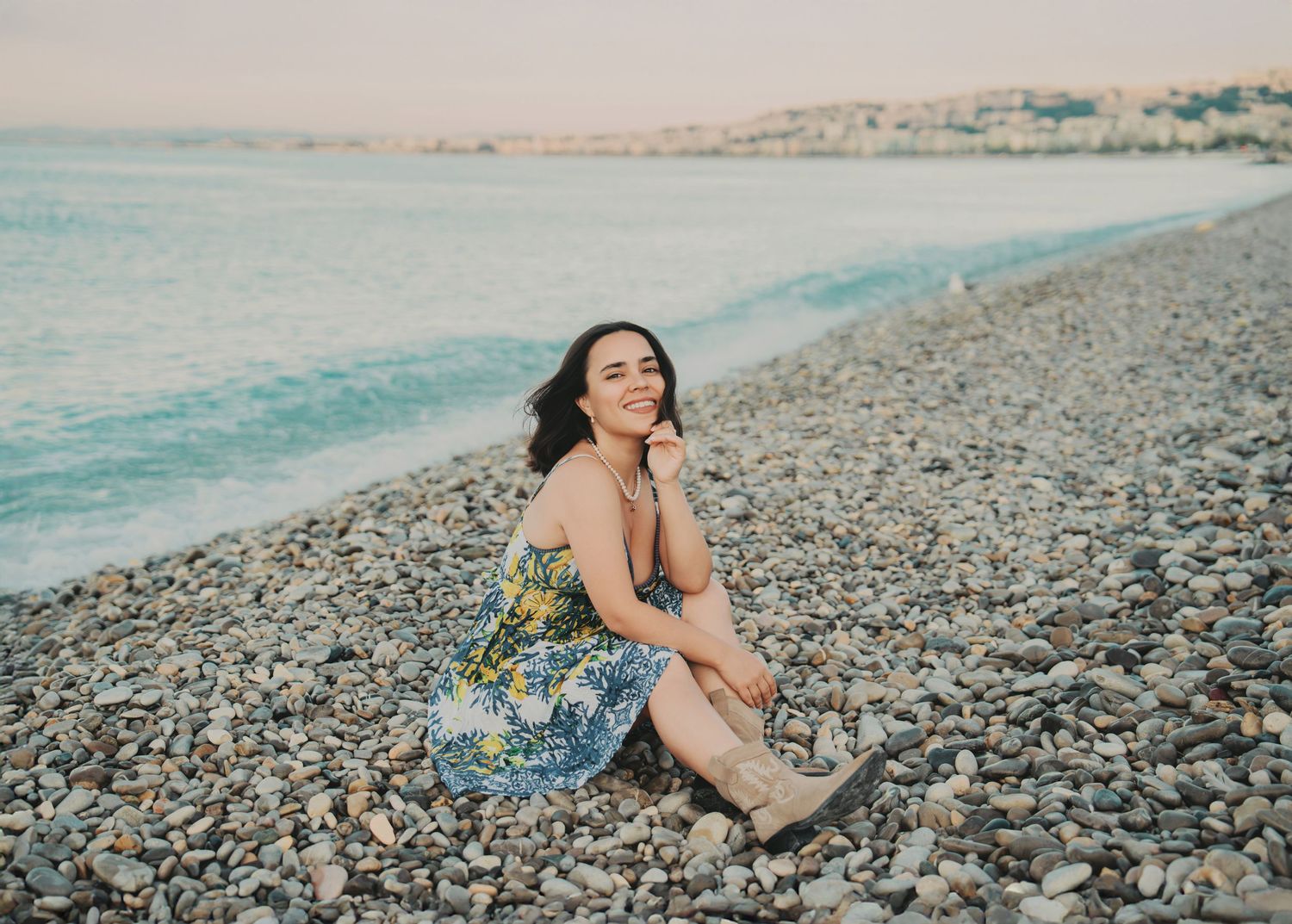 A happy woman in a floral dress sits laughing on a pebble beach at sunset.