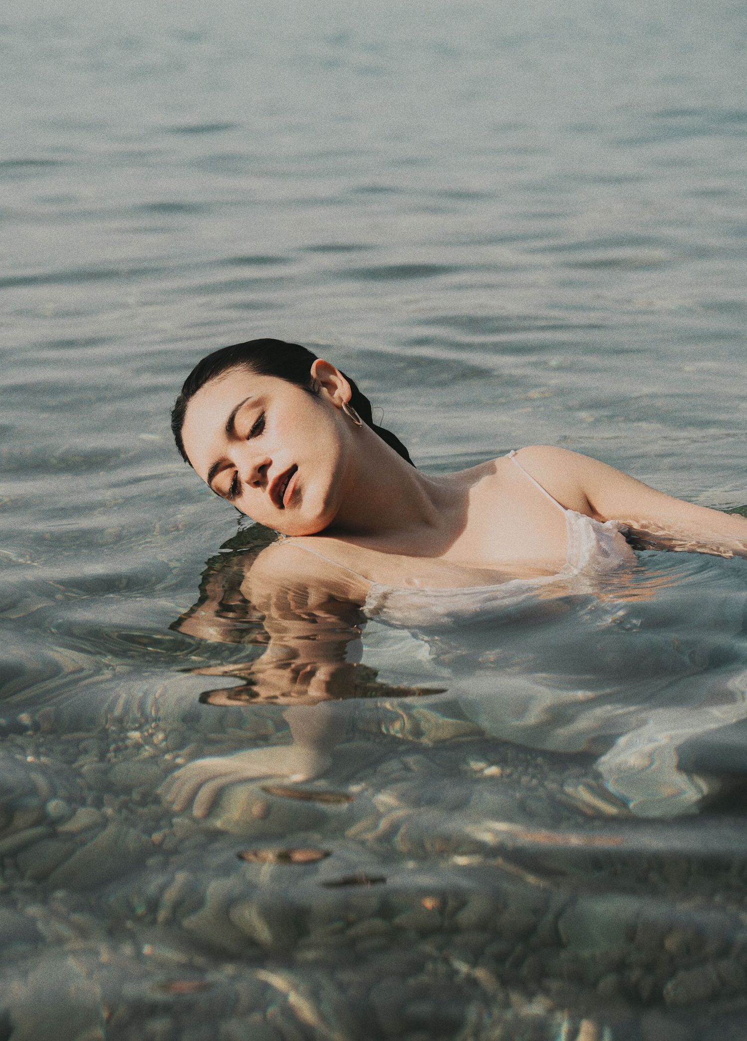 Young woman with dark hair floating in shallow clear water with eyes closed in serene pose.