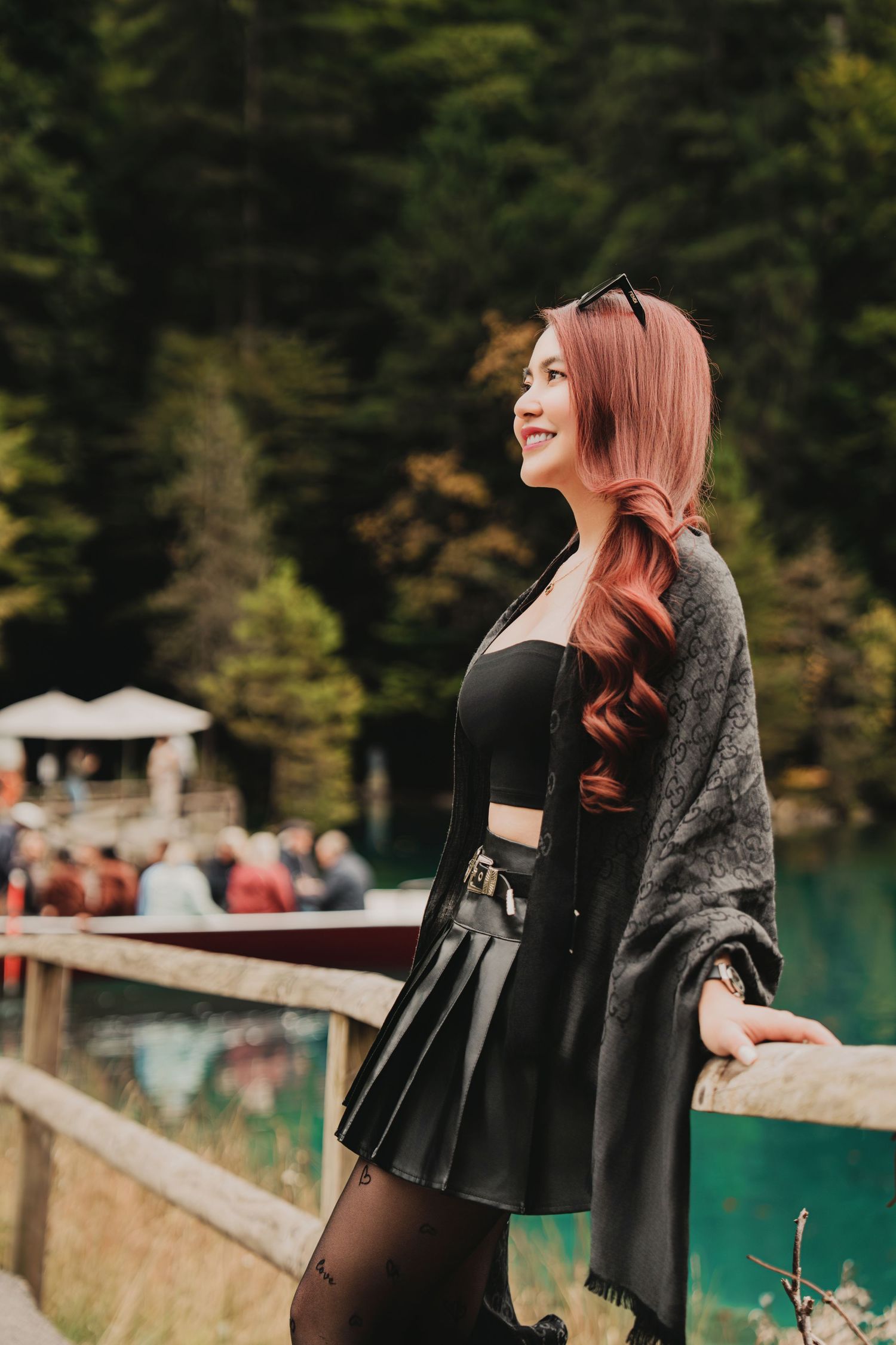 Cheerful red-haired woman in black skirt and gray shawl smiling beside turquoise alpine lake.