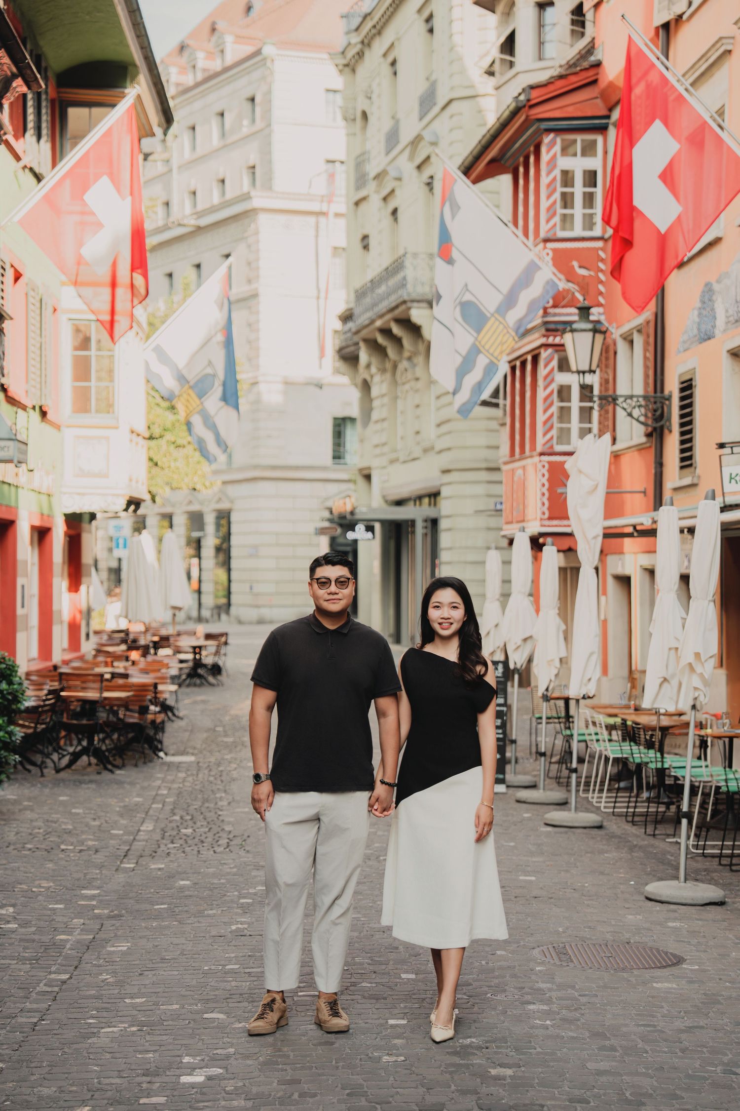 Couple holding hands on a Swiss cobblestone street lined with colorful buildings and Swiss flags.