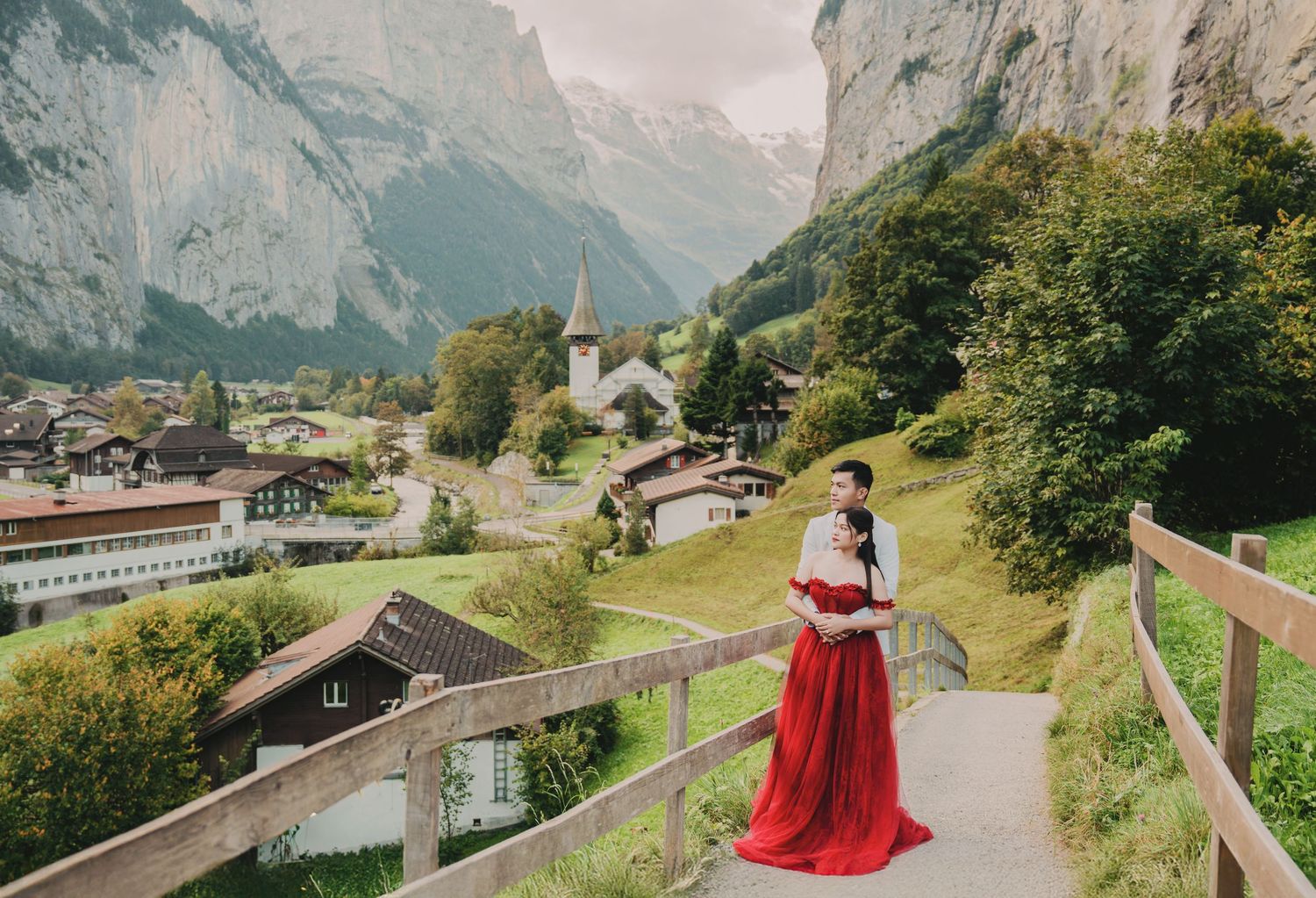 Romantic couple in red dress and white suit posing on wooden bridge in scenic Swiss mountain village.
