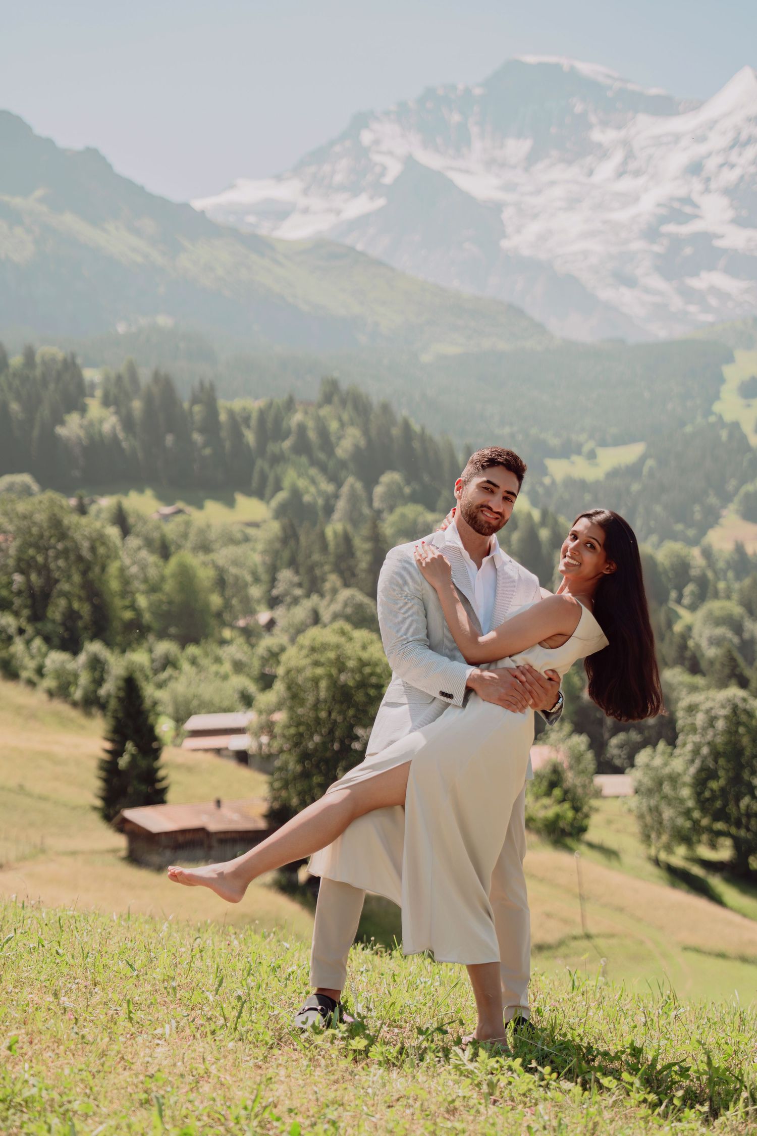 Groom lifting smiling bride on a green Alpine meadow with snow-capped mountains in the background.