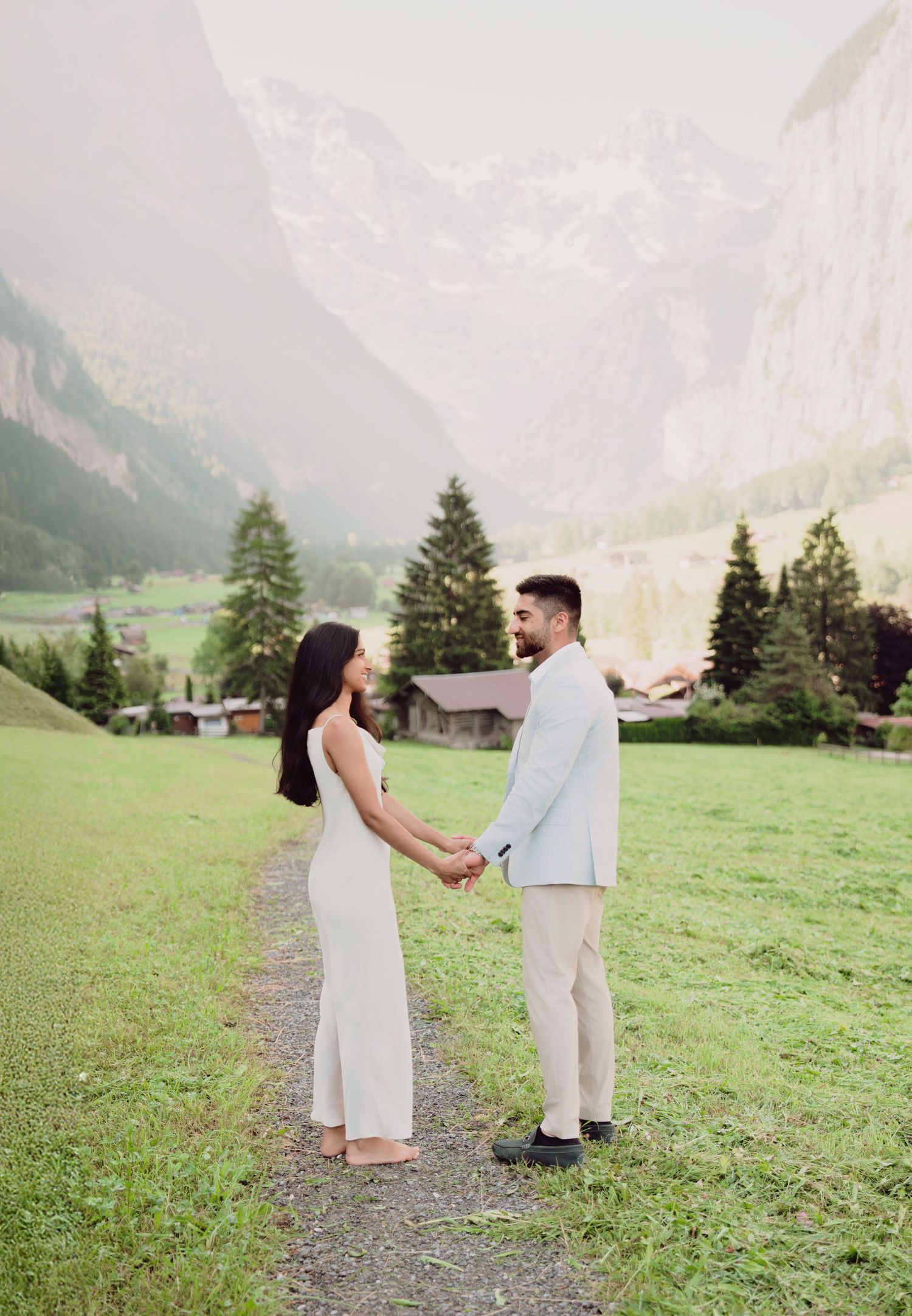 Bride and groom hold hands facing each other in a lush green valley surrounded by misty Swiss mountains.