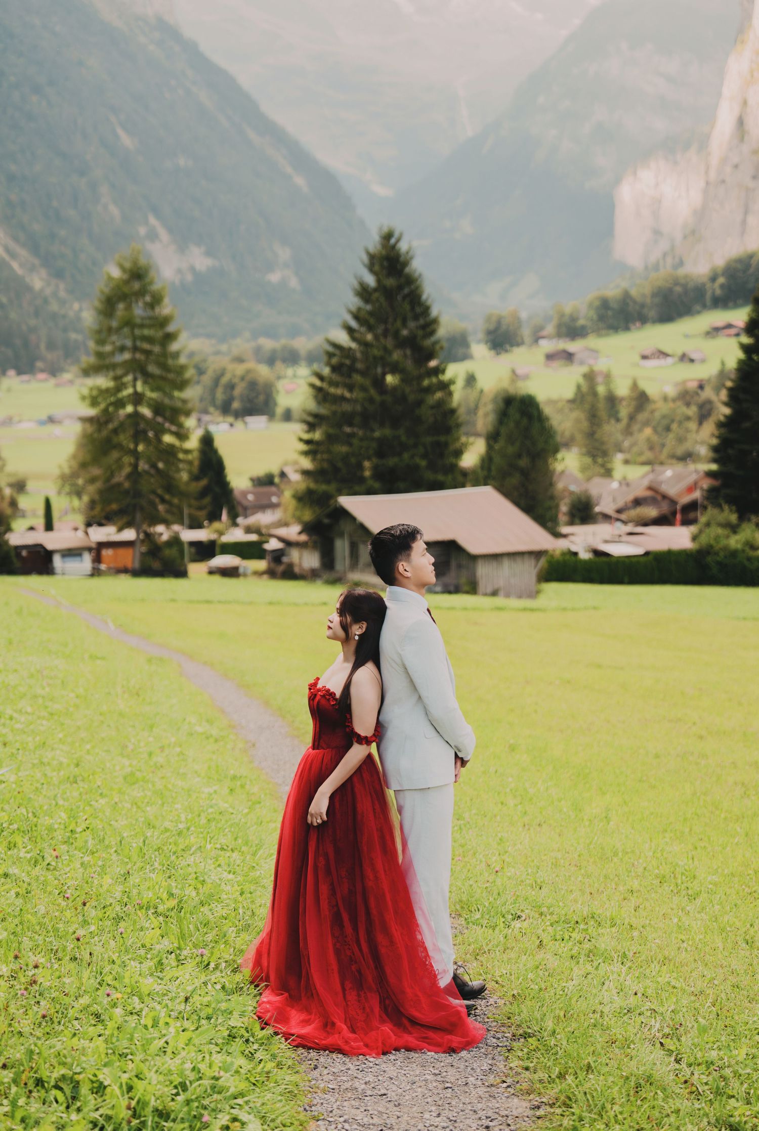 Couple standing back-to-back in Swiss alpine meadow, bride in red gown, groom in grey suit.