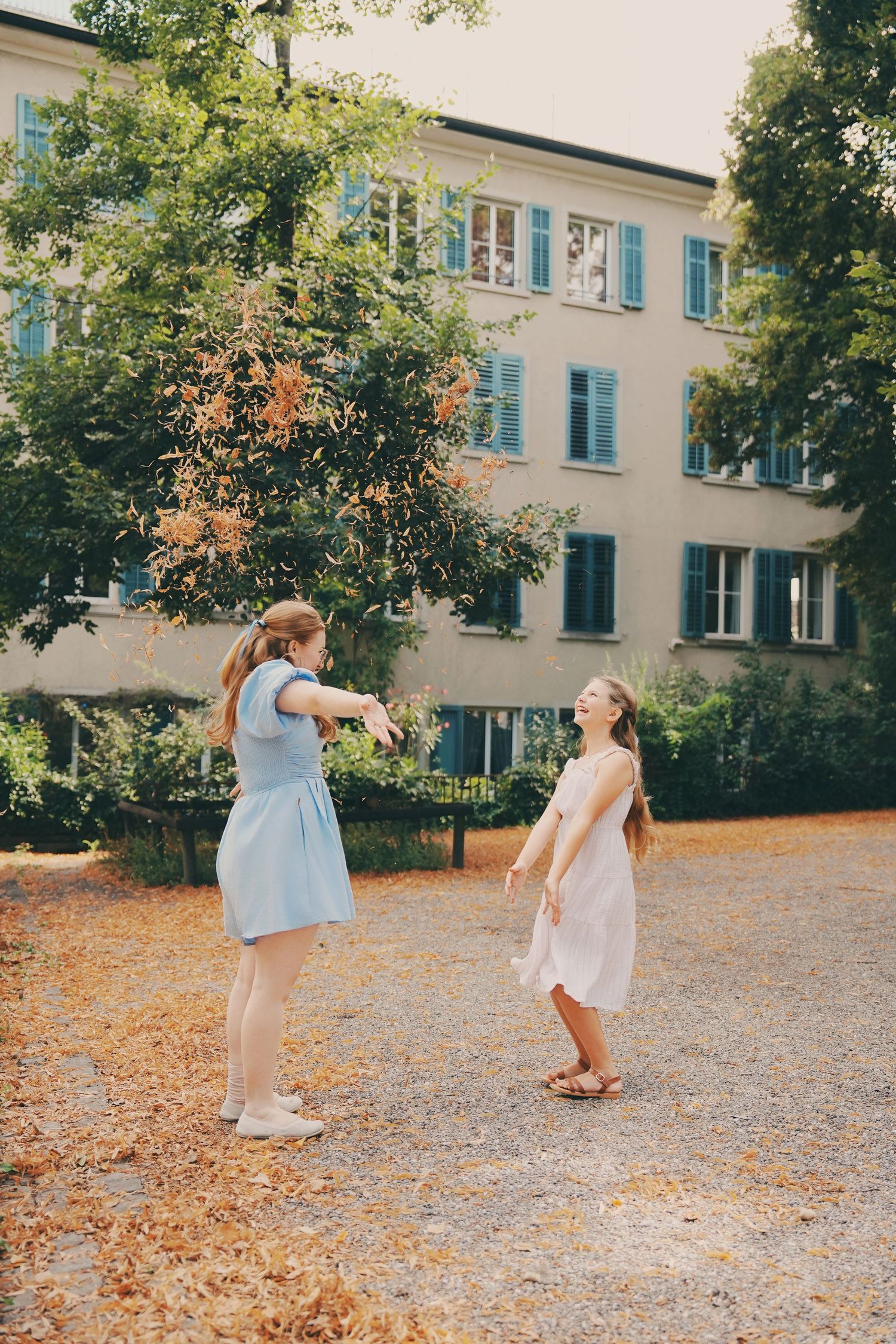 Two girls in summer dresses play outdoors in front of a European building surrounded by trees.