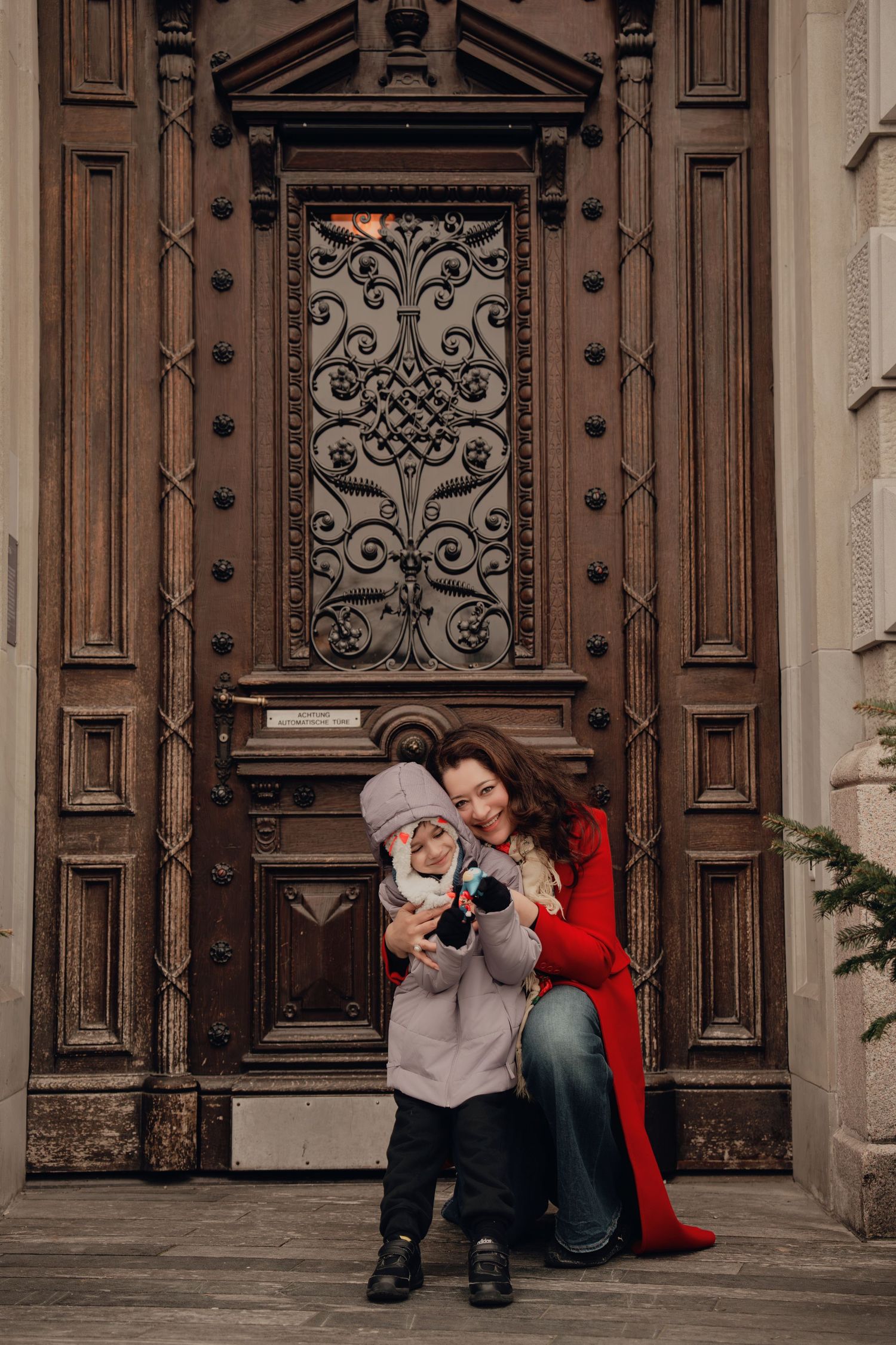 Mother in red coat kneeling beside young child holding a camera at decorative wooden door.