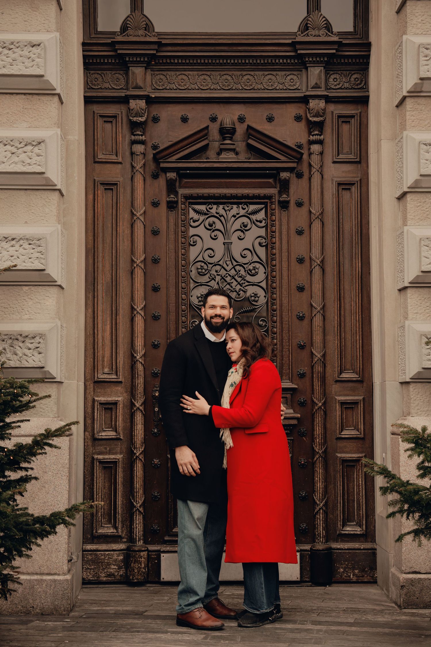 Couple embracing romantically before an ornate carved wooden door of a historic building.