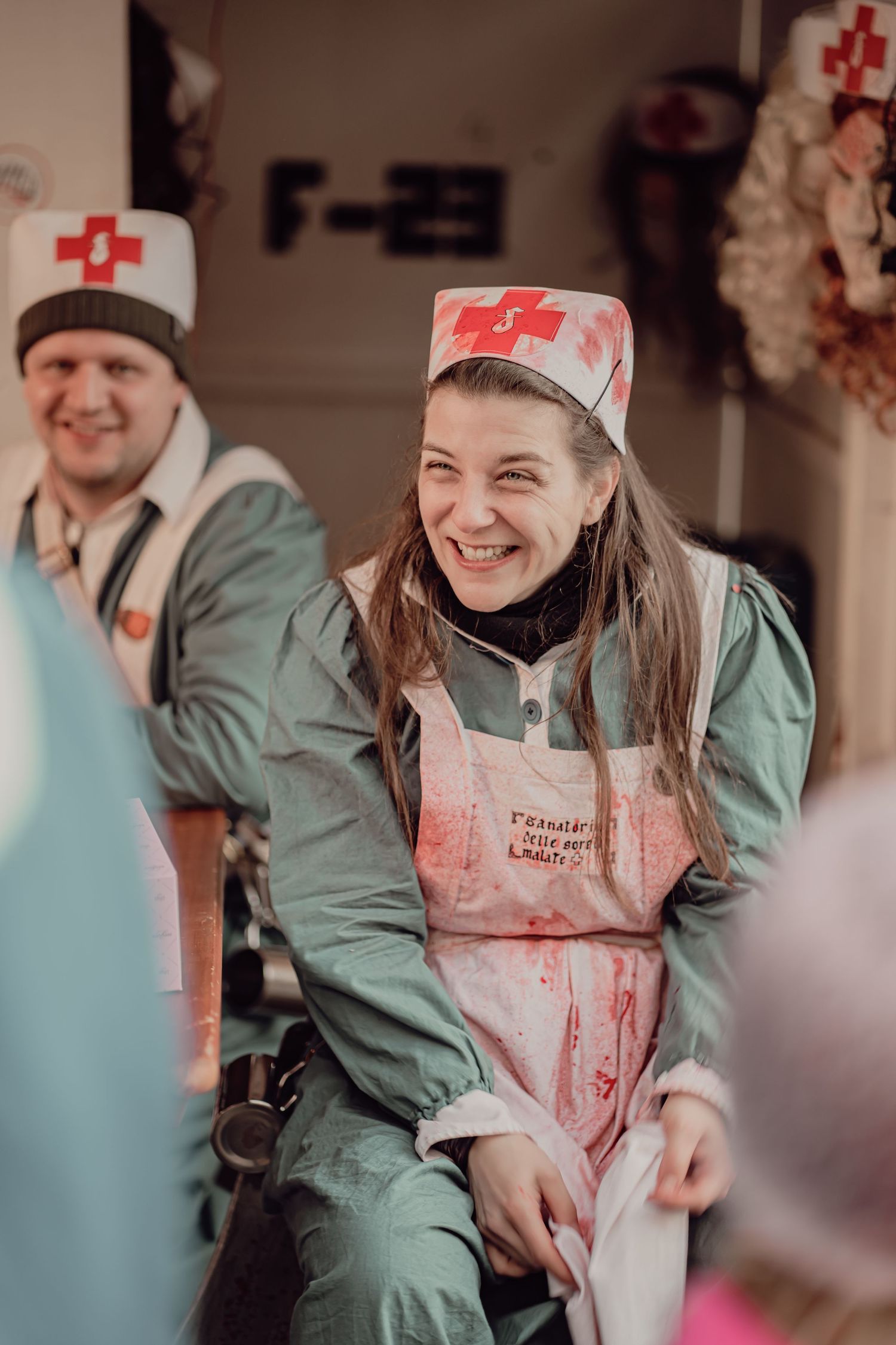 Laughing woman in bloody nurse Halloween costume sits at a festive carnival event.