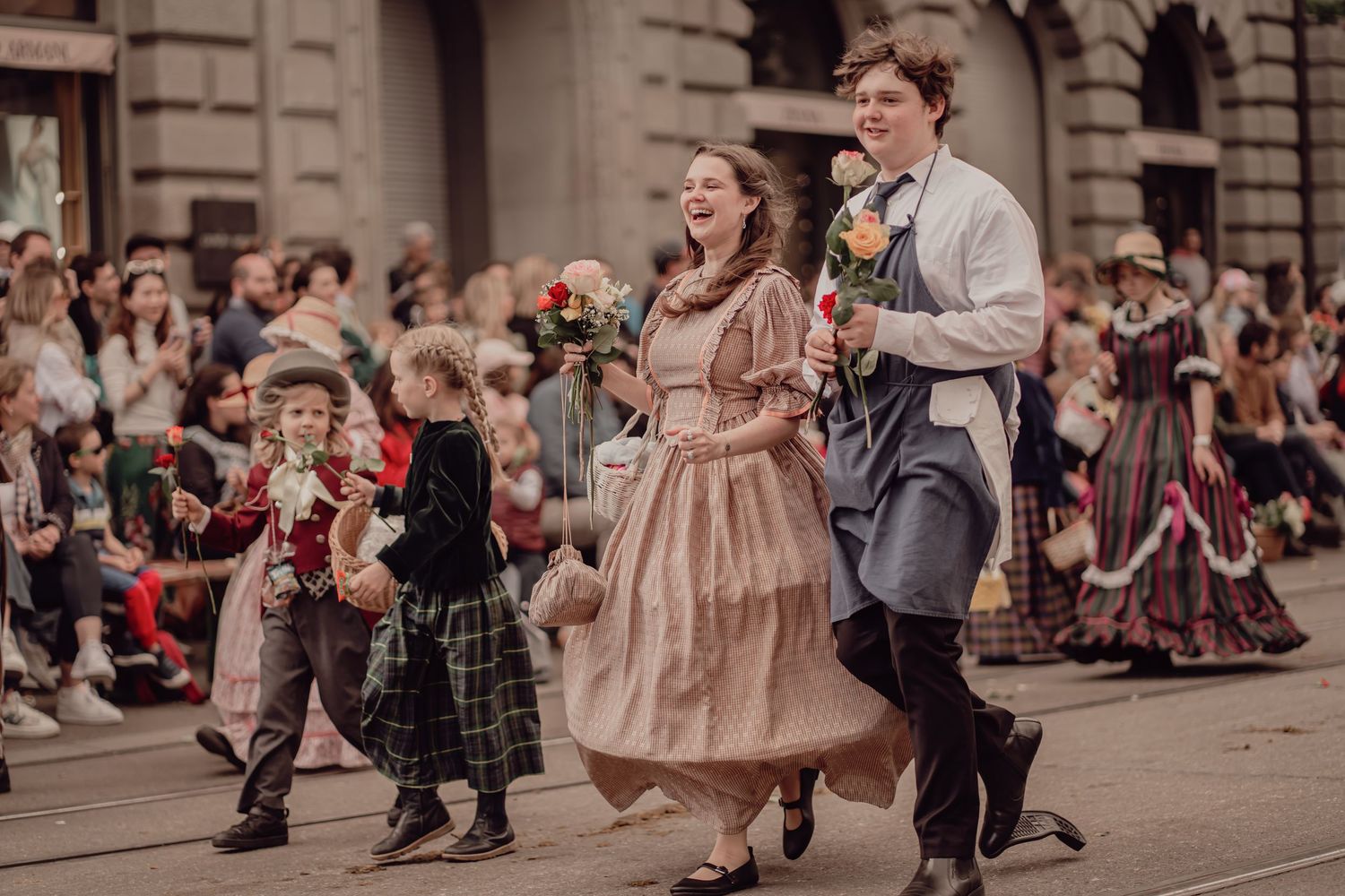 People in Victorian costumes parade through city streets, with a laughing woman in pink dress holding flowers.