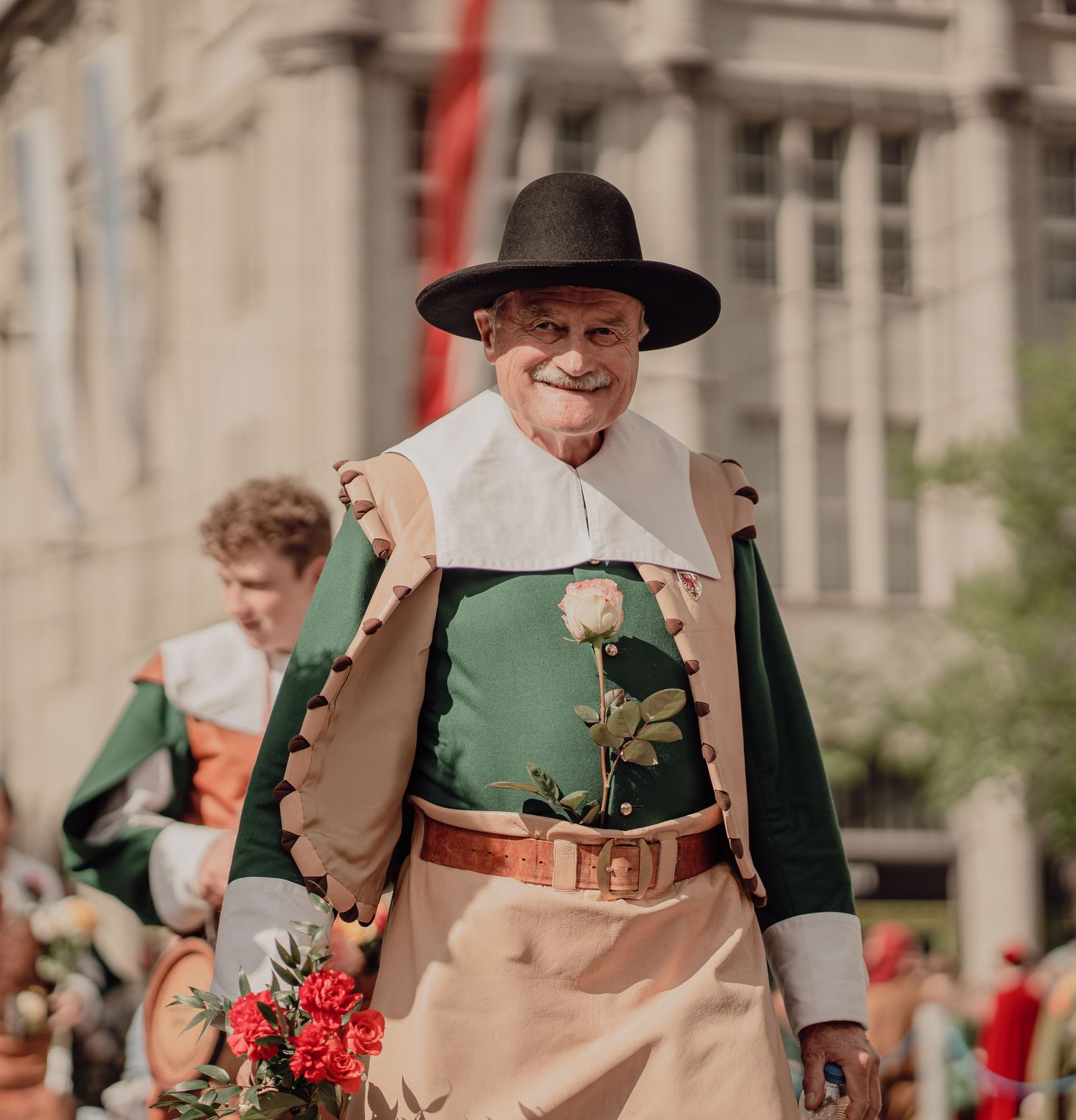 Smiling elderly man in green and cream Renaissance costume holds a white rose in a historical parade.