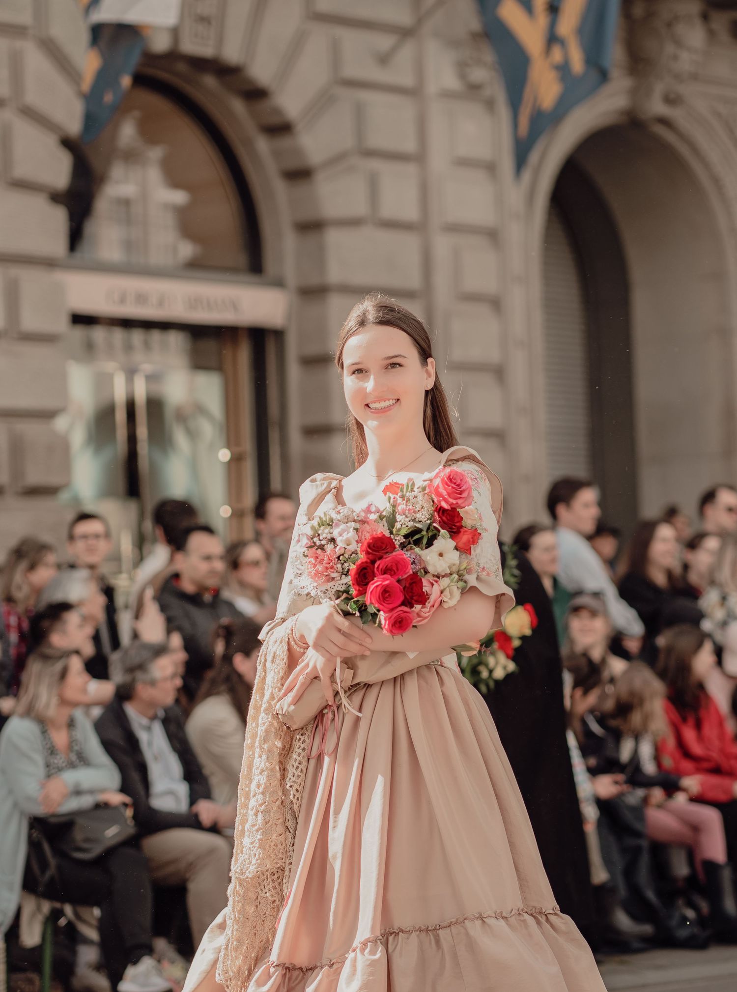 Young woman in elegant pink gown holding bouquet of roses at outdoor fashion event.