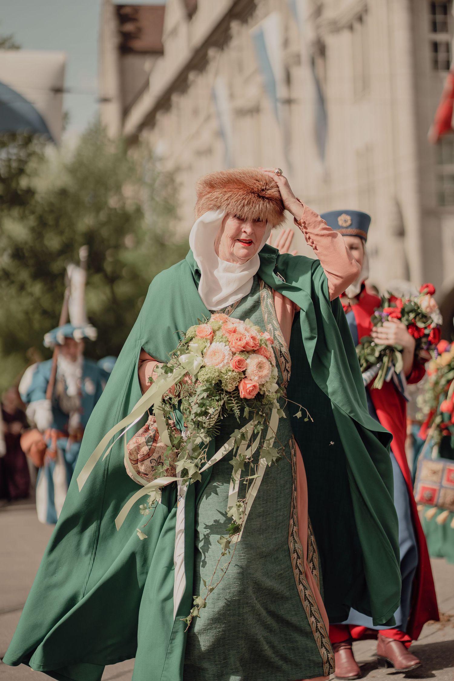 Woman in green cape and fur hat holds bouquet at traditional festival parade.