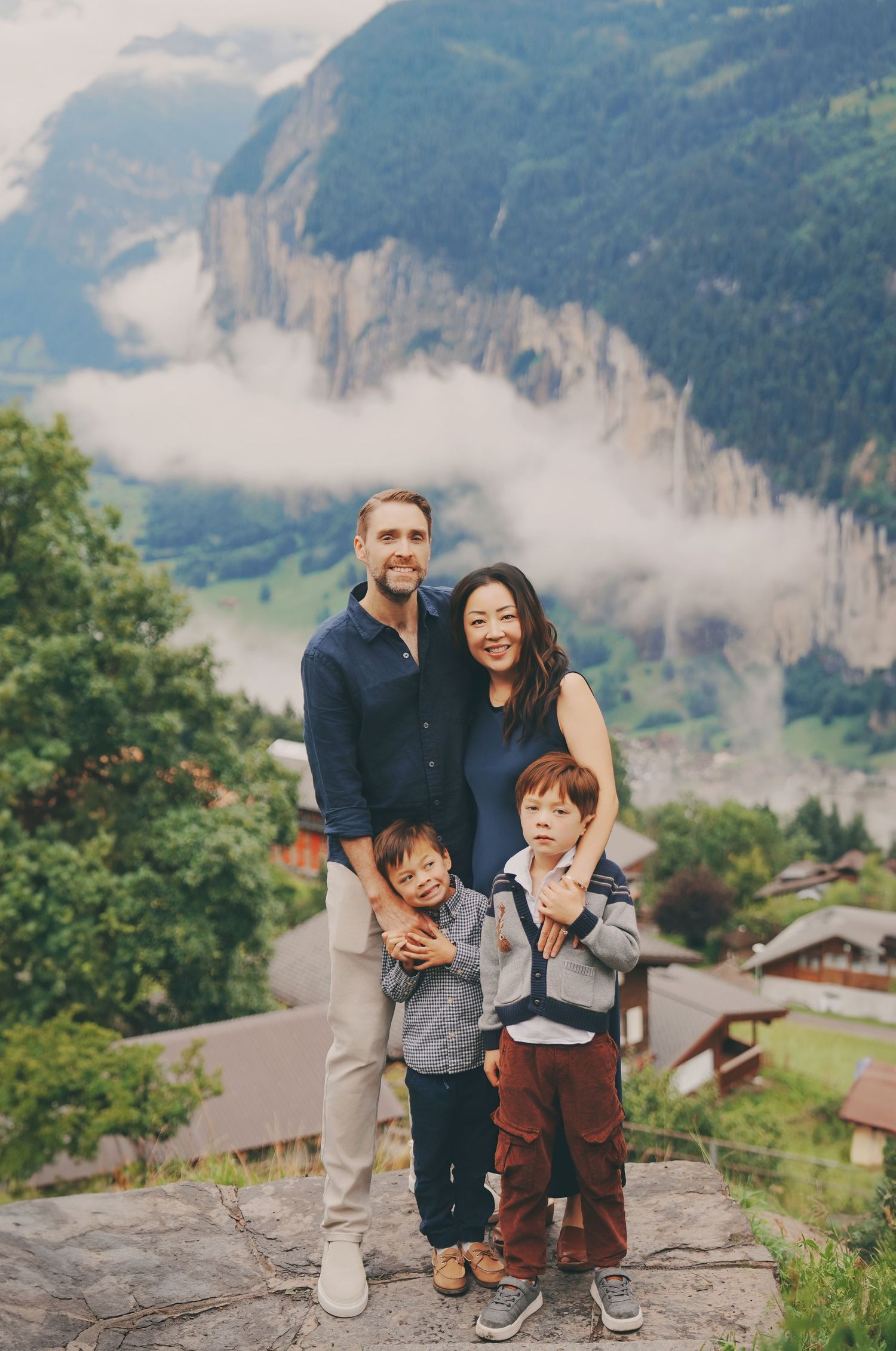 Family of four poses on rocky overlook with dramatic Swiss Alps, clouds, and village below.
