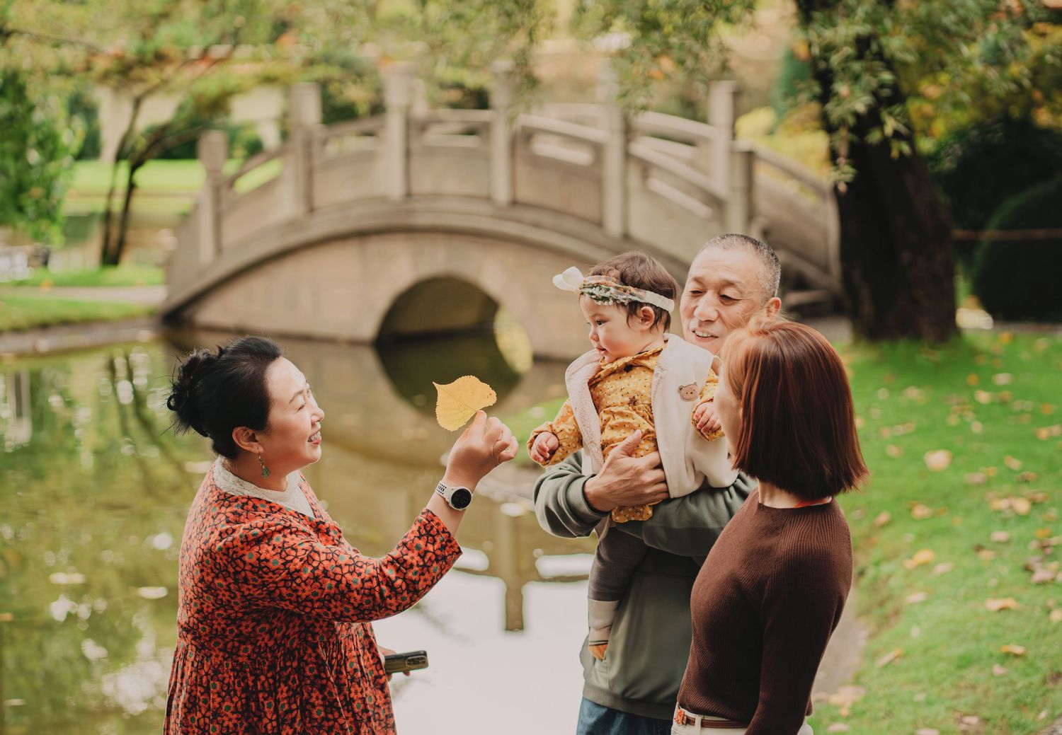 Multigenerational Asian family sharing red envelopes with baby near stone bridge in autumn park.