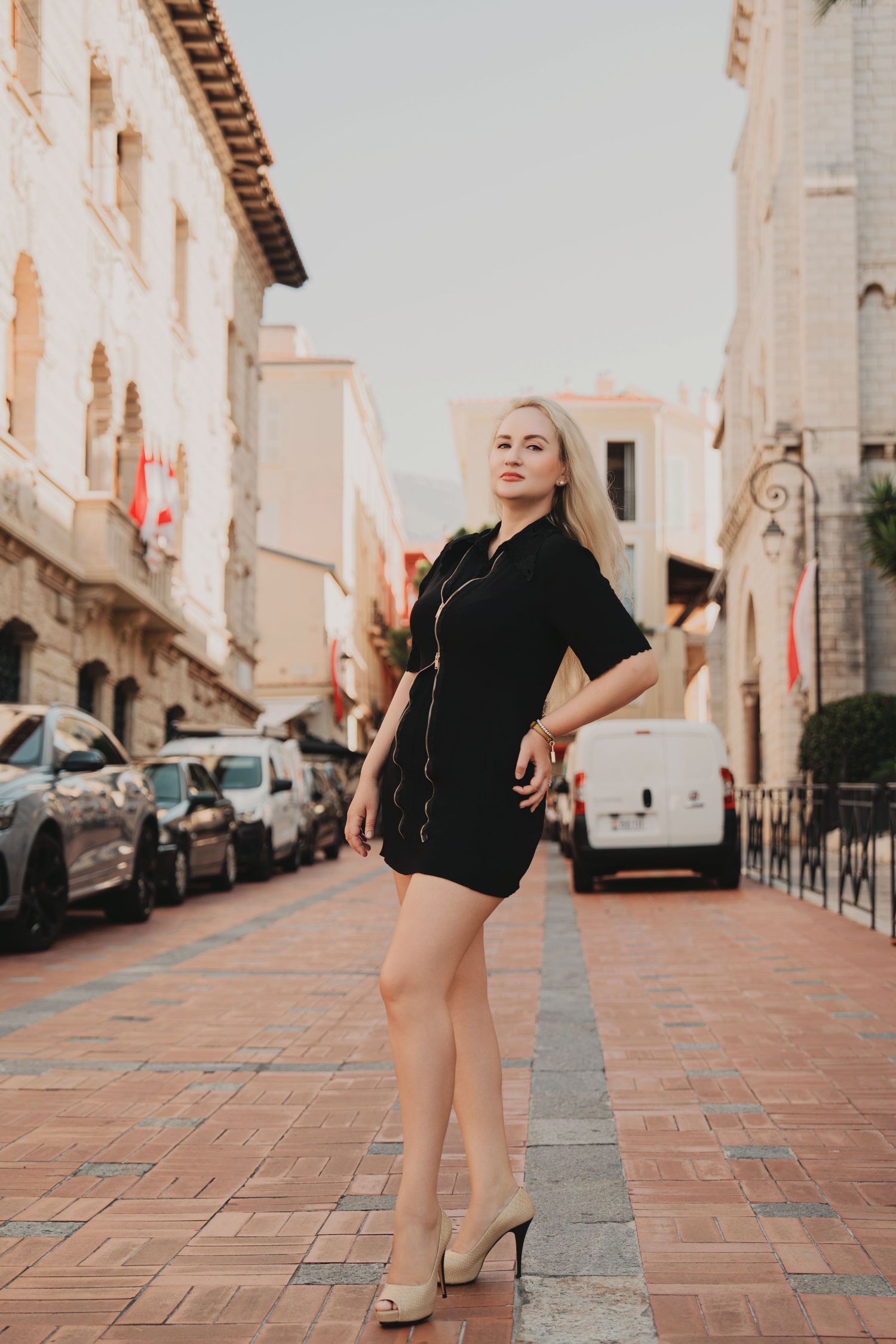 Elegant blonde woman in black dress and heels standing on a cobblestone European street.