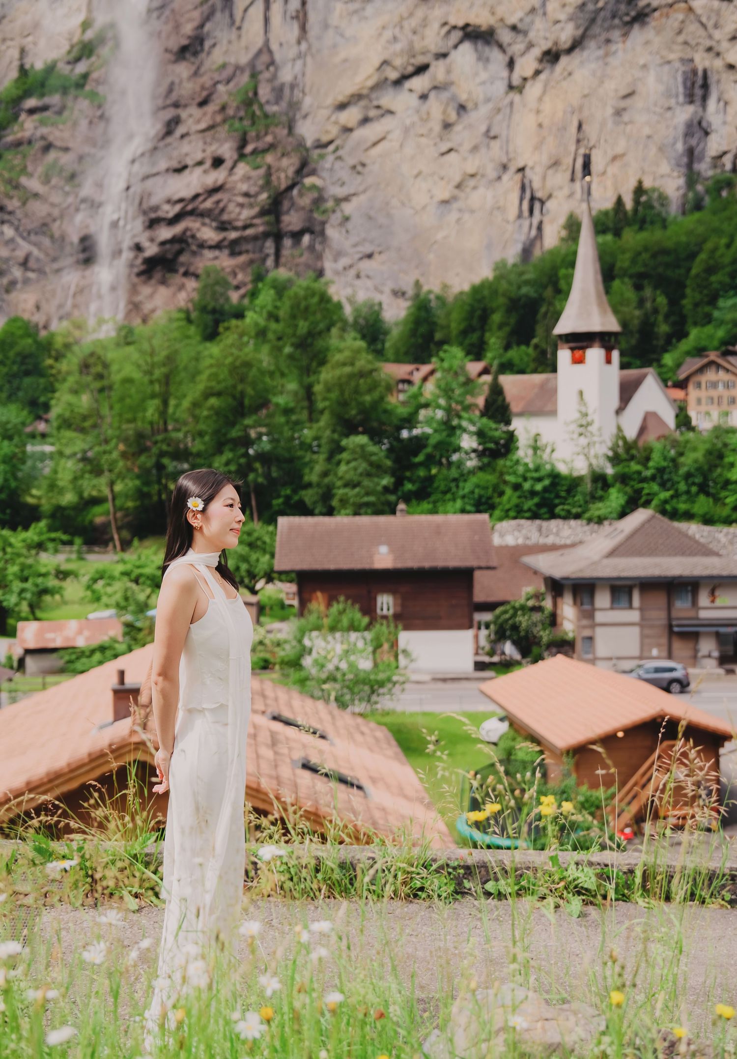 Woman in a white dress standing near wildflowers with Staubbach Falls and a church steeple in Lauterbrunnen.