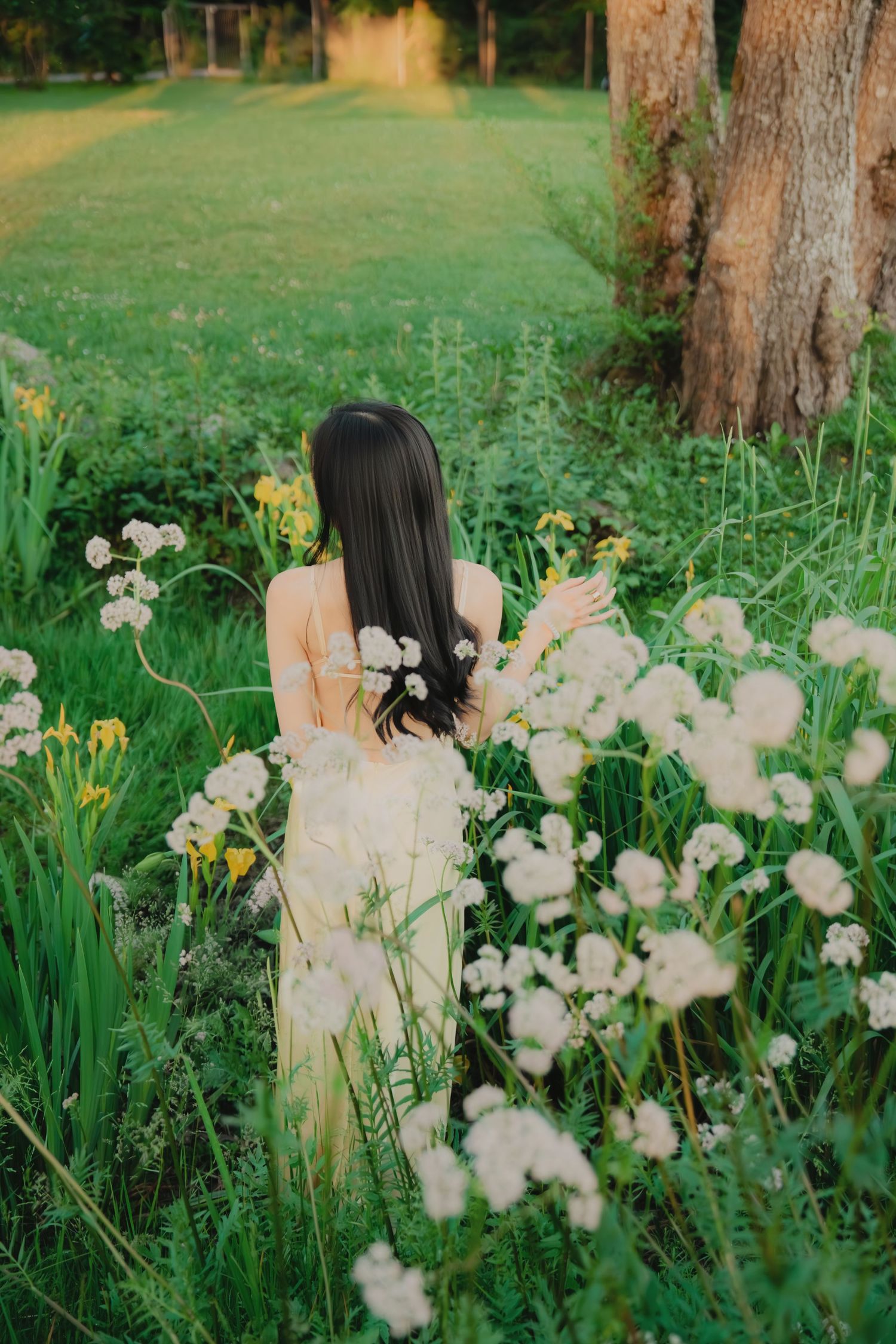 Woman in yellow dress with flowers in hair standing among white wildflowers in lush green garden.