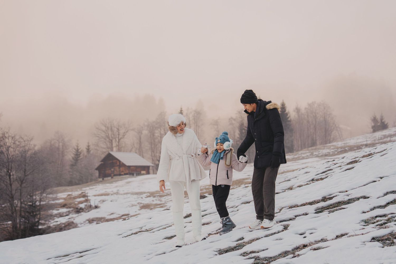 Family of three walking on snowy hillside in winter, wearing warm coats with misty mountains and wooden cabin in background.