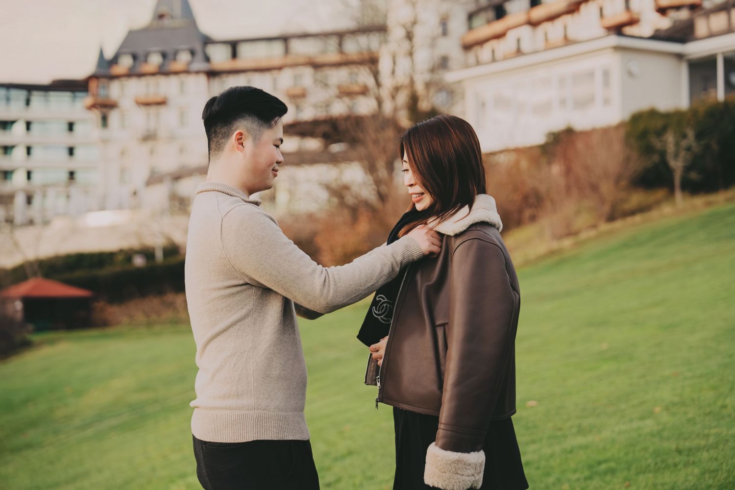 Young Asian man adjusting woman's jacket collar on green lawn outside hotel.