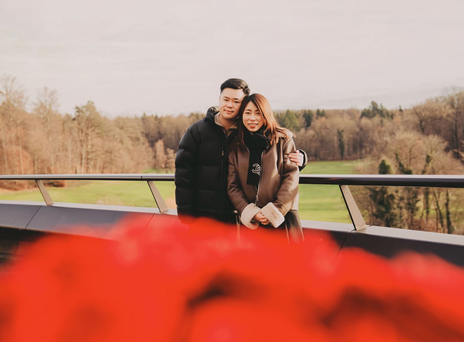 Happy Asian couple embracing on a balcony with lush green landscape and red flowers in foreground.