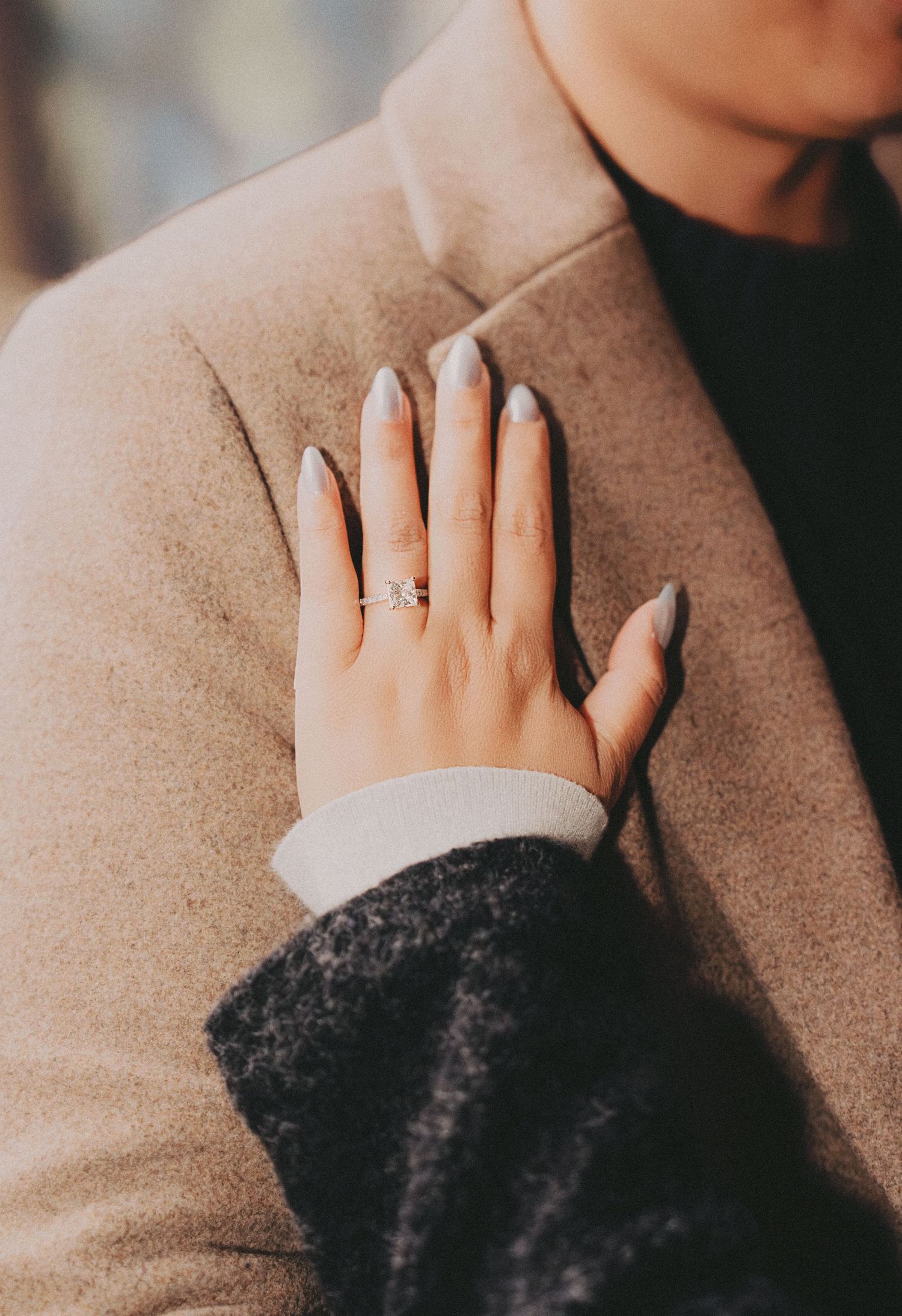 Close-up of a woman's hand displaying a diamond engagement ring on a beige wool coat.