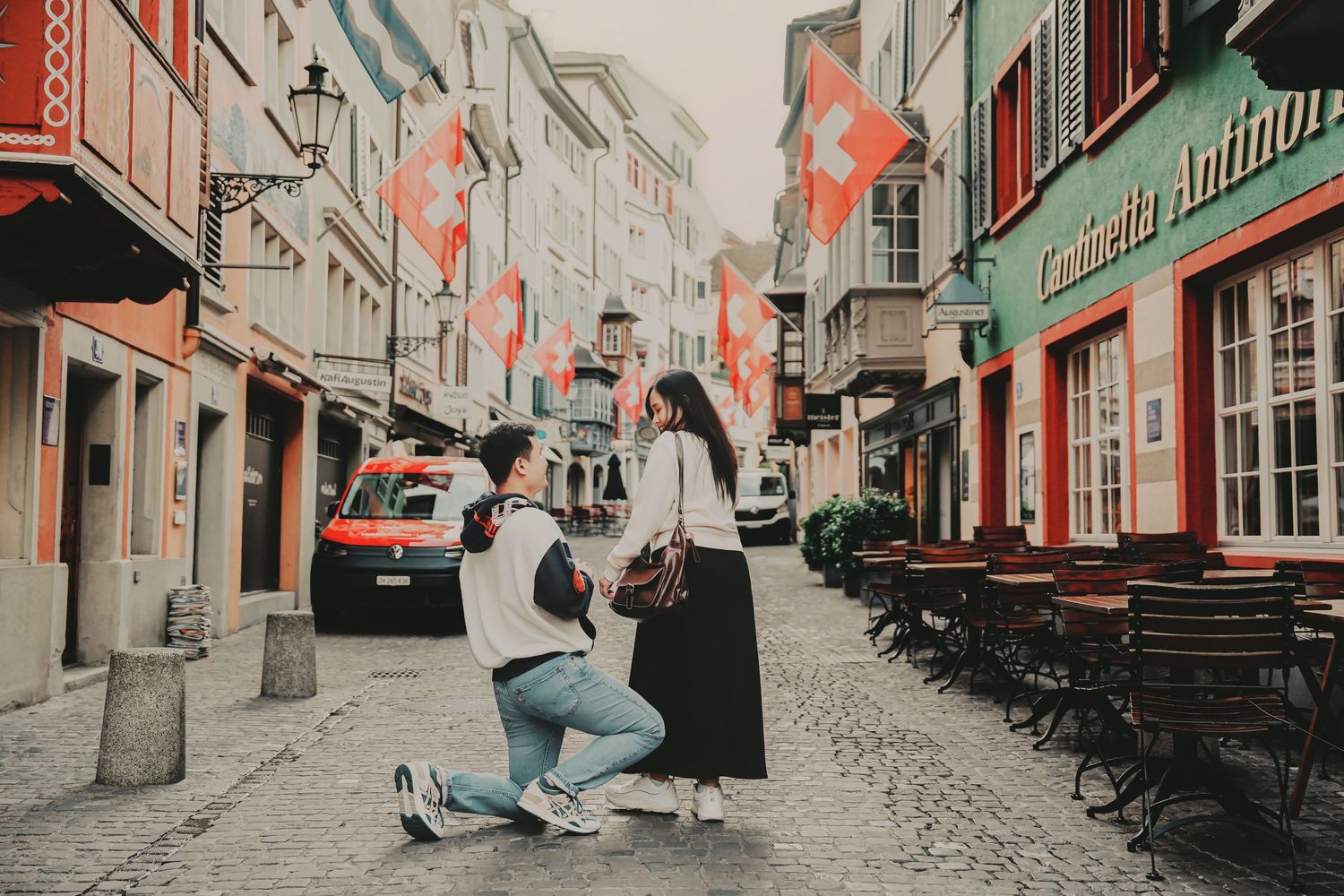 Man kneeling to propose to woman on a charming Swiss cobblestone street lined with red Swiss flags.