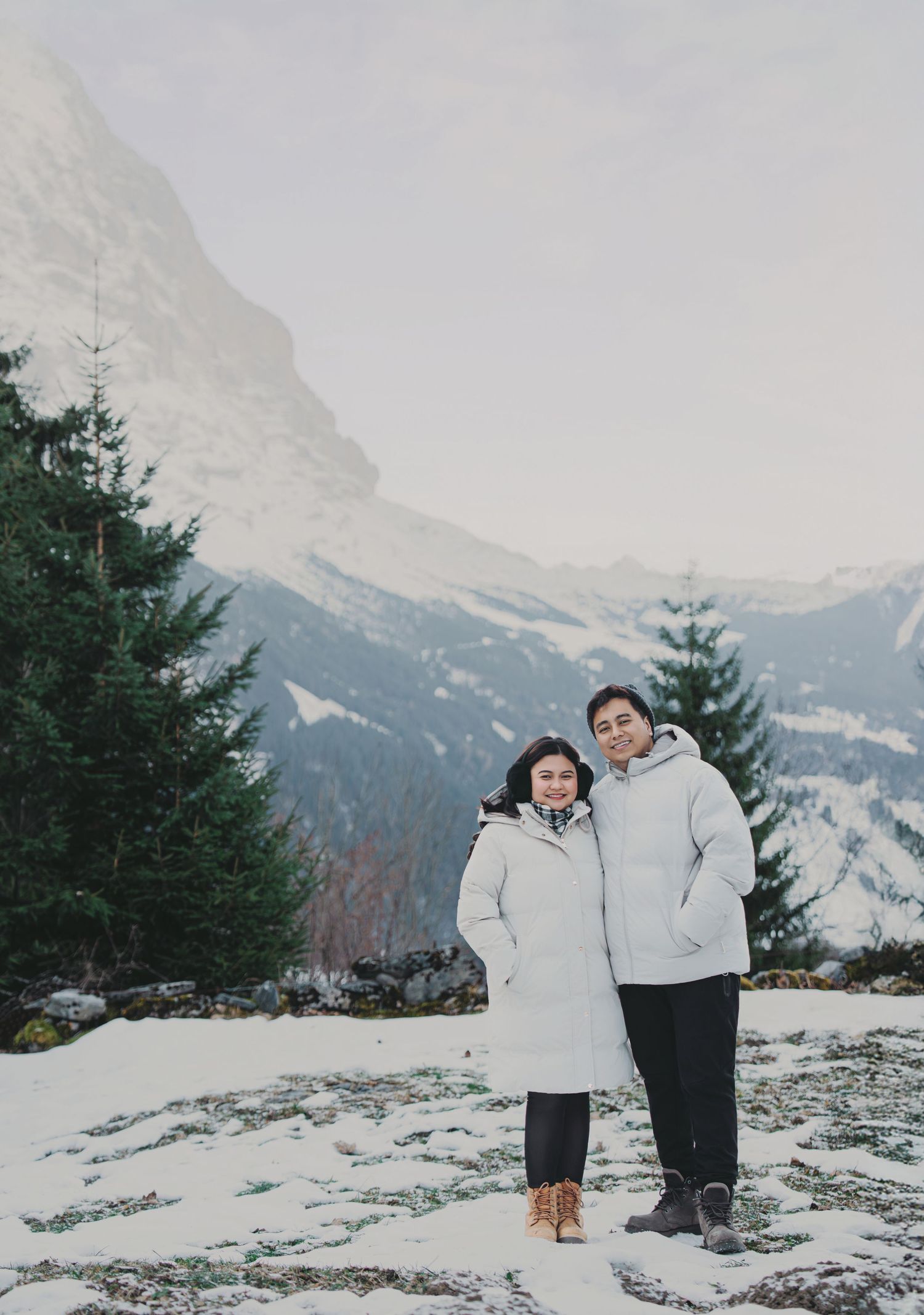 Couple in matching white puffer coats posing together in snowy alpine landscape with mountain peak.