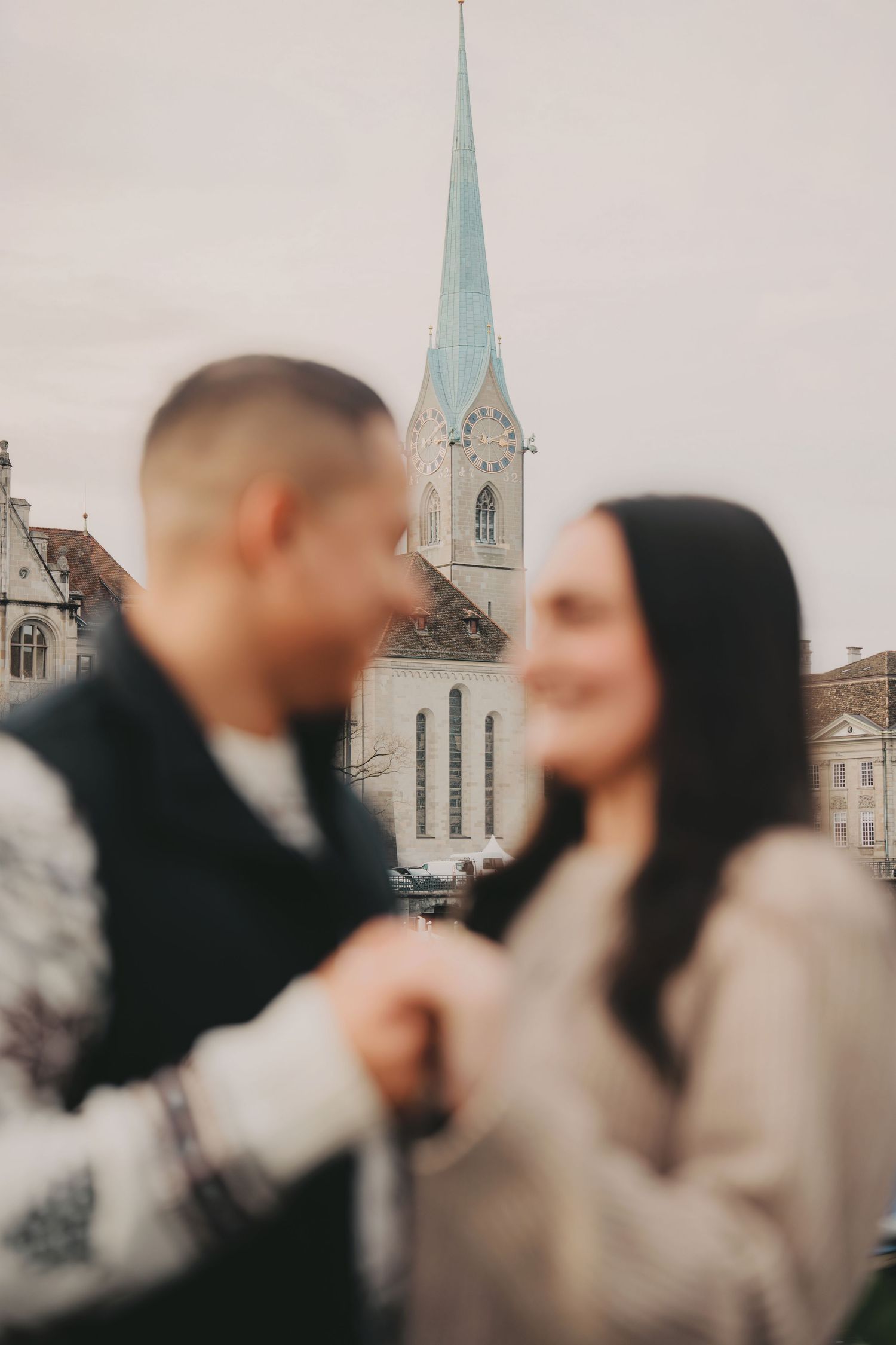 Blurred couple holding hands on a bridge with Fraumunster Church spire in Zurich background.